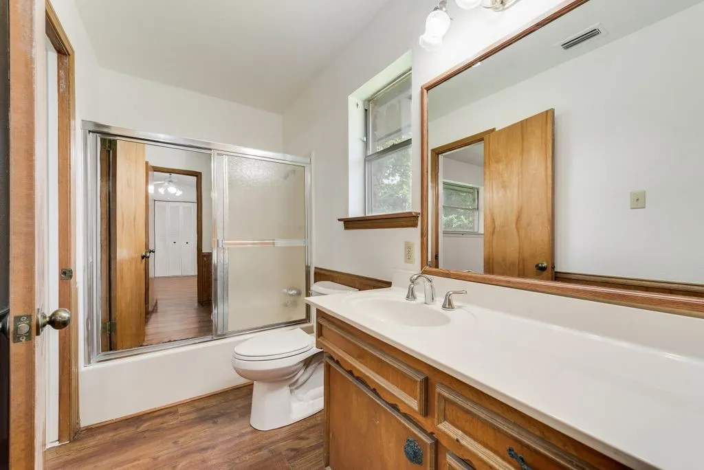 Bathroom featuring wood-finish flooring, a white vanity with integrated sink, a large framed mirror, a tub-shower combination with sliding glass doors, and a single window