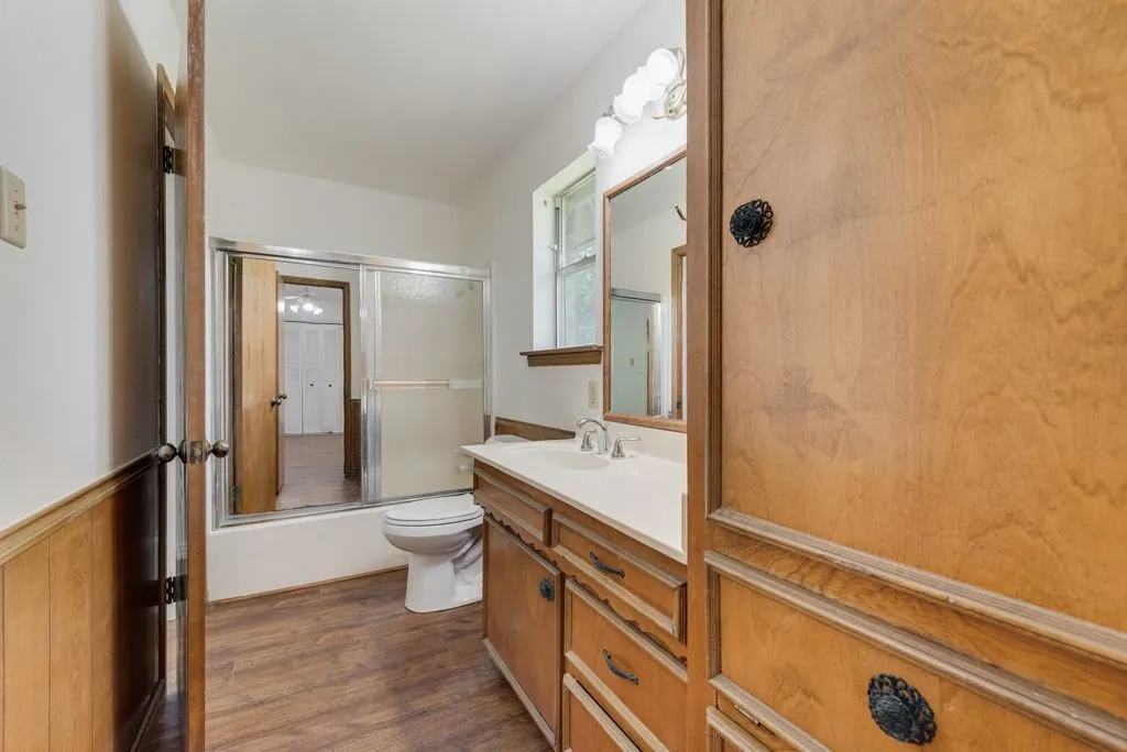 Full bathroom featuring wood-finish flooring, a wood vanity with a white countertop and integrated sink, a mirrored medicine cabinet, a full bathtub with a sliding glass enclosure, and a built-in wood cabinet with decorative hardware