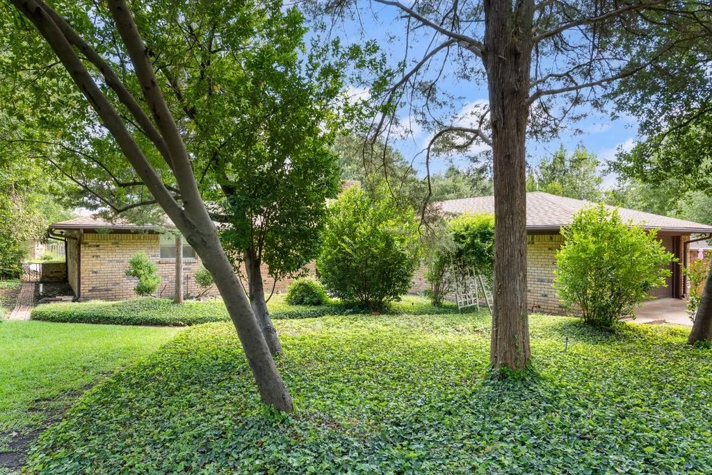 Brick exterior home featuring a brown shingle roof, mature trees, manicured shrubs, and a lush ground cover