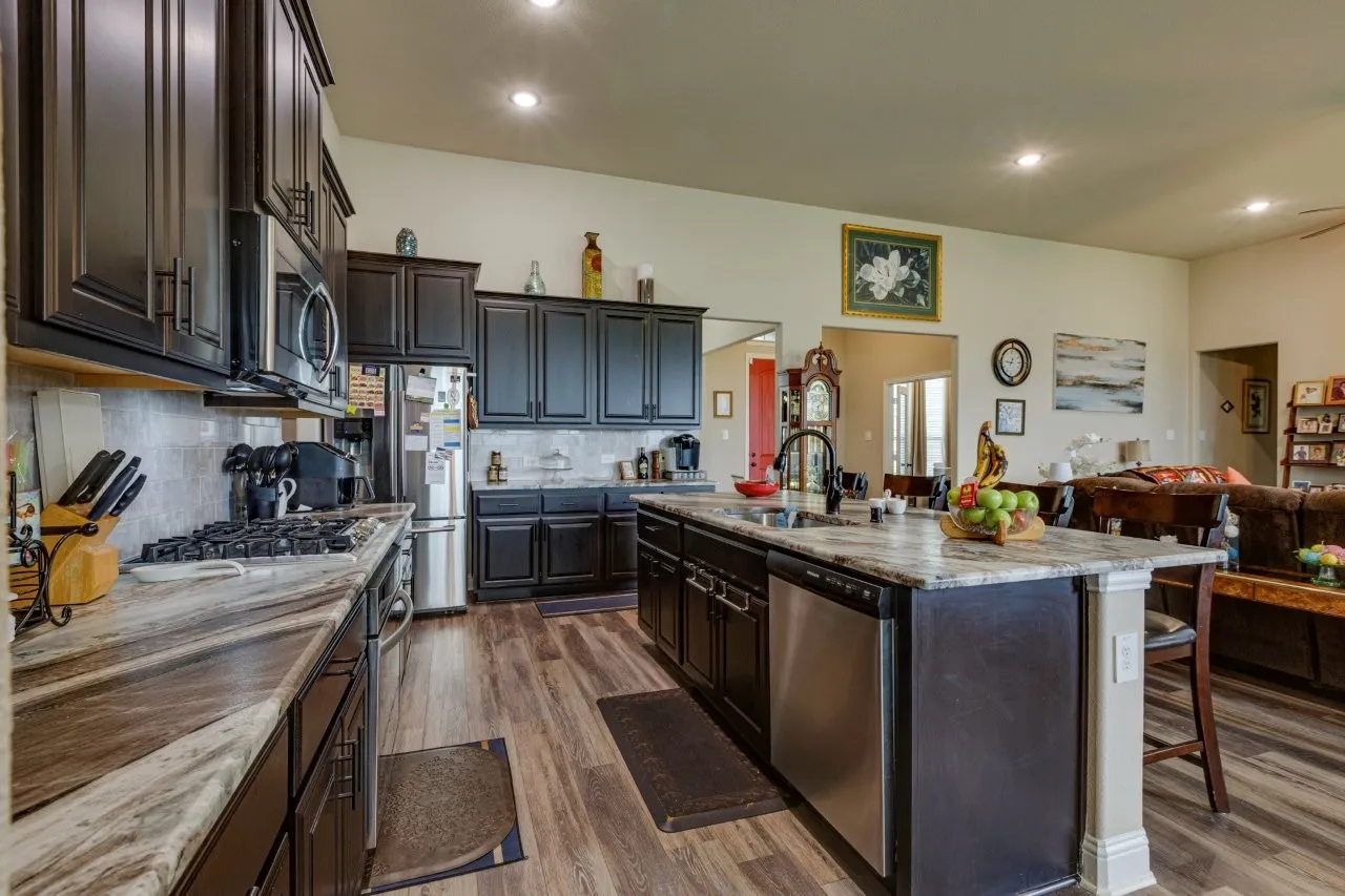 Spacious kitchen featuring dark wood cabinetry, stainless steel appliances, variegated countertops, a kitchen island with integrated sink, and wood-finish flooring