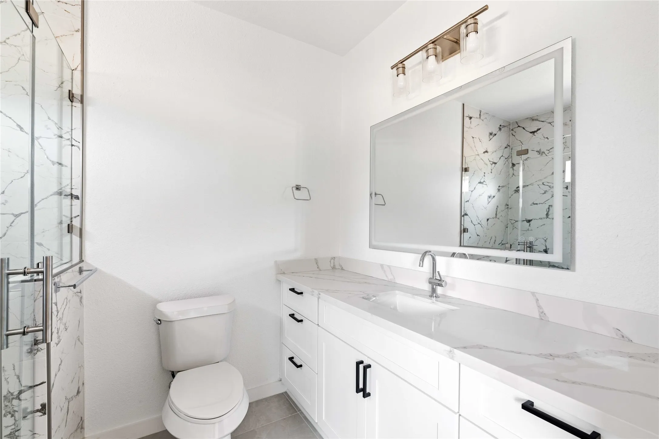 White vanity with integrated sink and chrome faucet, rectangular framed mirror, three-light vanity bar, glass-enclosed shower with marble-look tile, and grey tile flooring