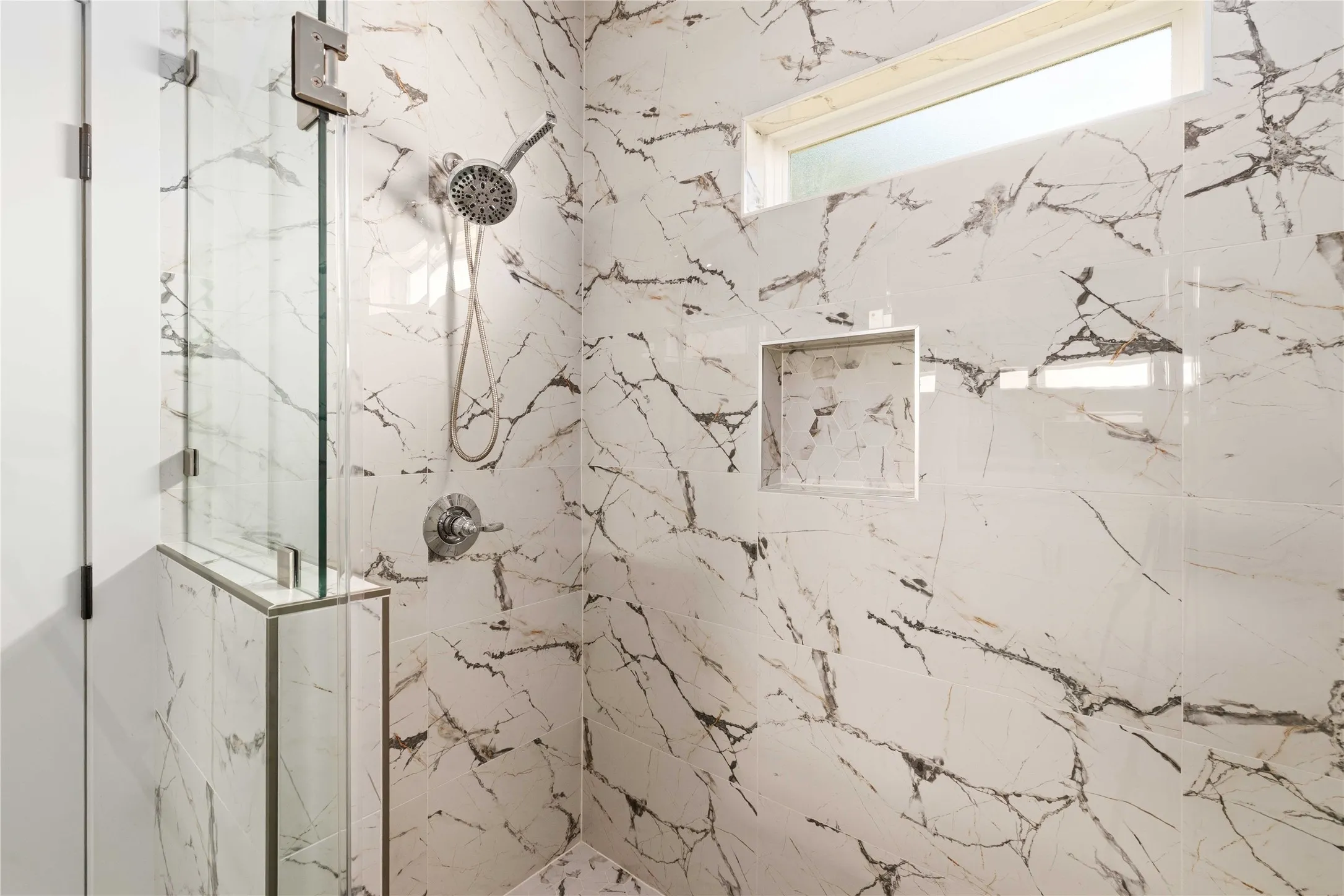 Glass-enclosed shower with a handheld shower fixture, built-in niche, and marble-patterned tile walls and flooring