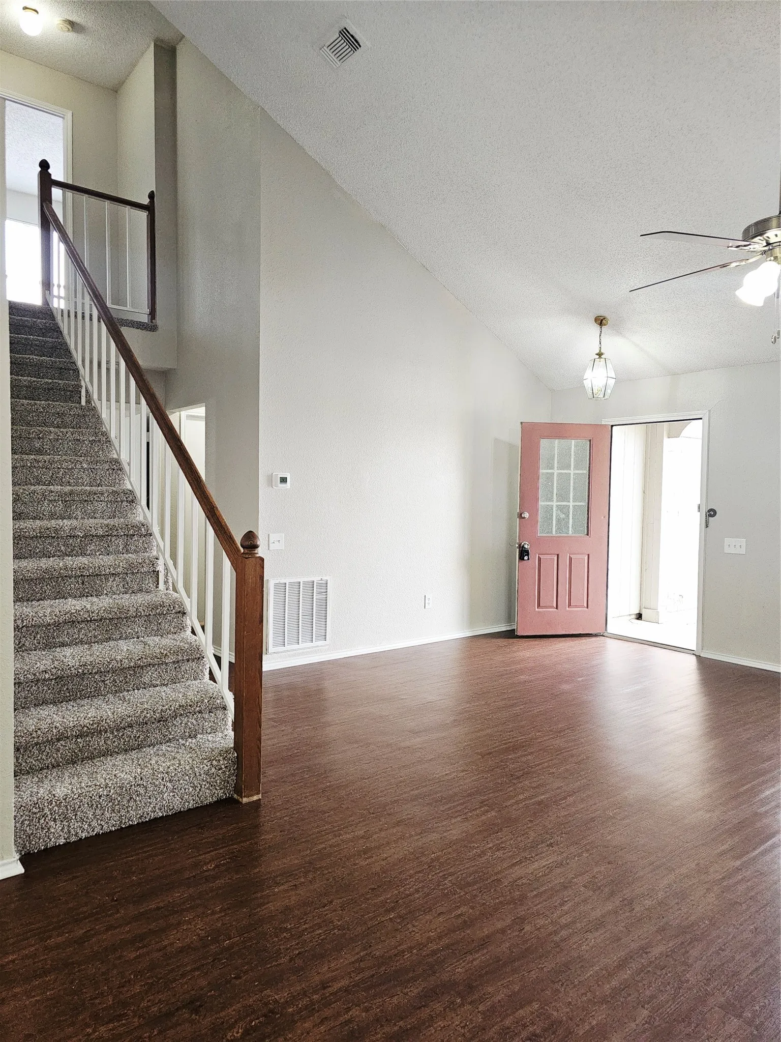 Inviting entryway featuring a pink paneled door with glass inserts, wood-finish flooring, a carpeted staircase with wood banister, a vaulted ceiling, and a ceiling fan with integrated lighting