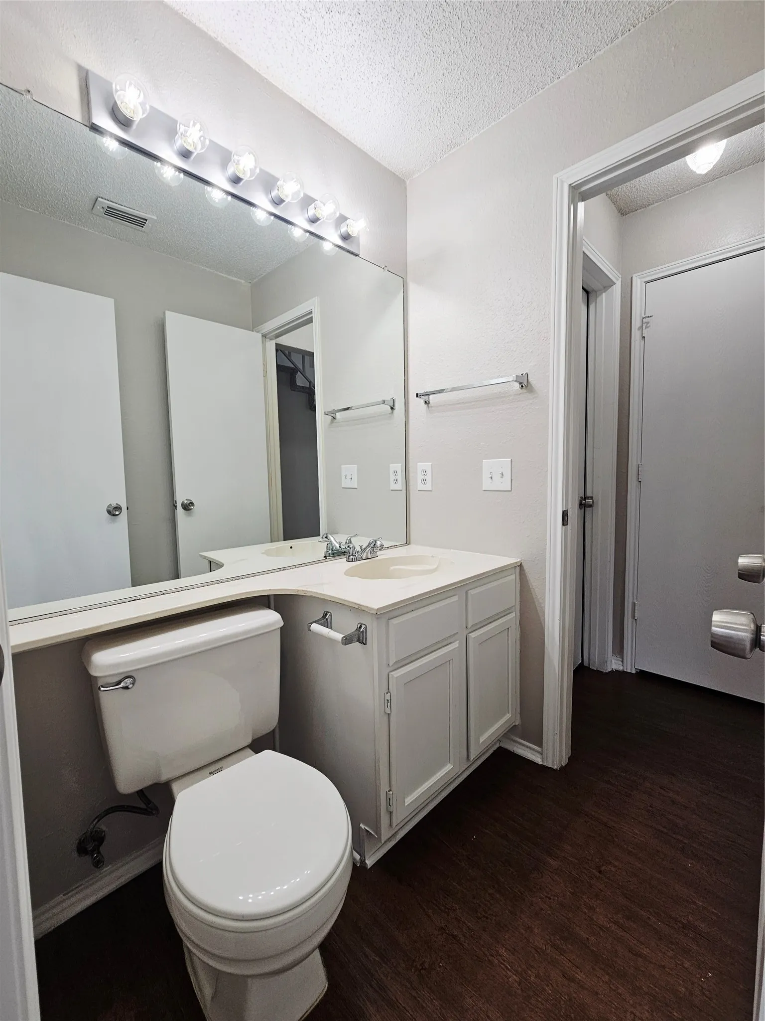Bathroom featuring a vanity with a white cabinet and countertop, a large wall-mounted mirror, a strip light fixture with exposed bulbs, and dark wood-finish flooring