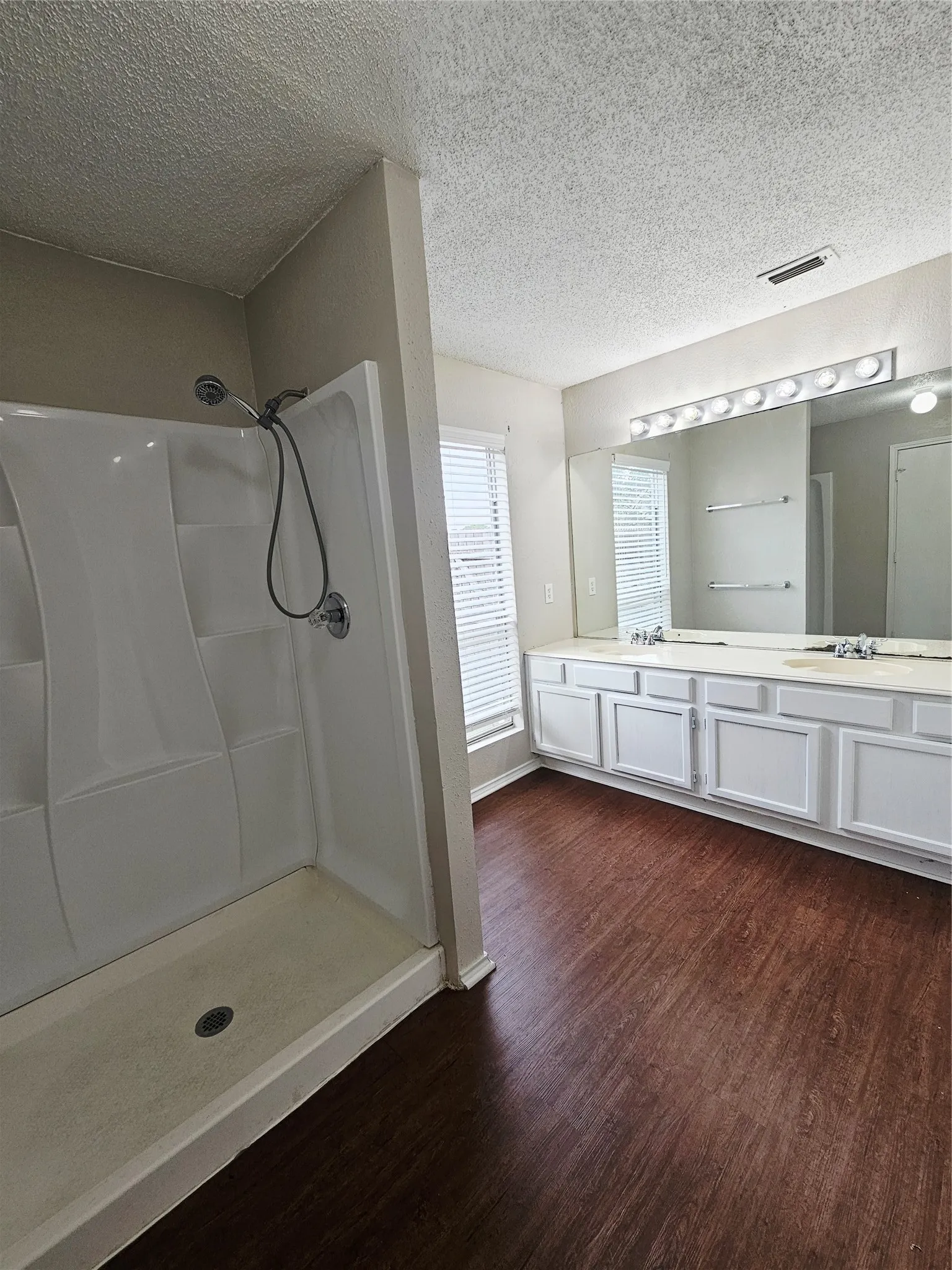 Bathroom featuring a walk-in shower with chrome fixtures, wood-finish flooring, a double vanity with white cabinetry, and horizontal blinds on the windows
