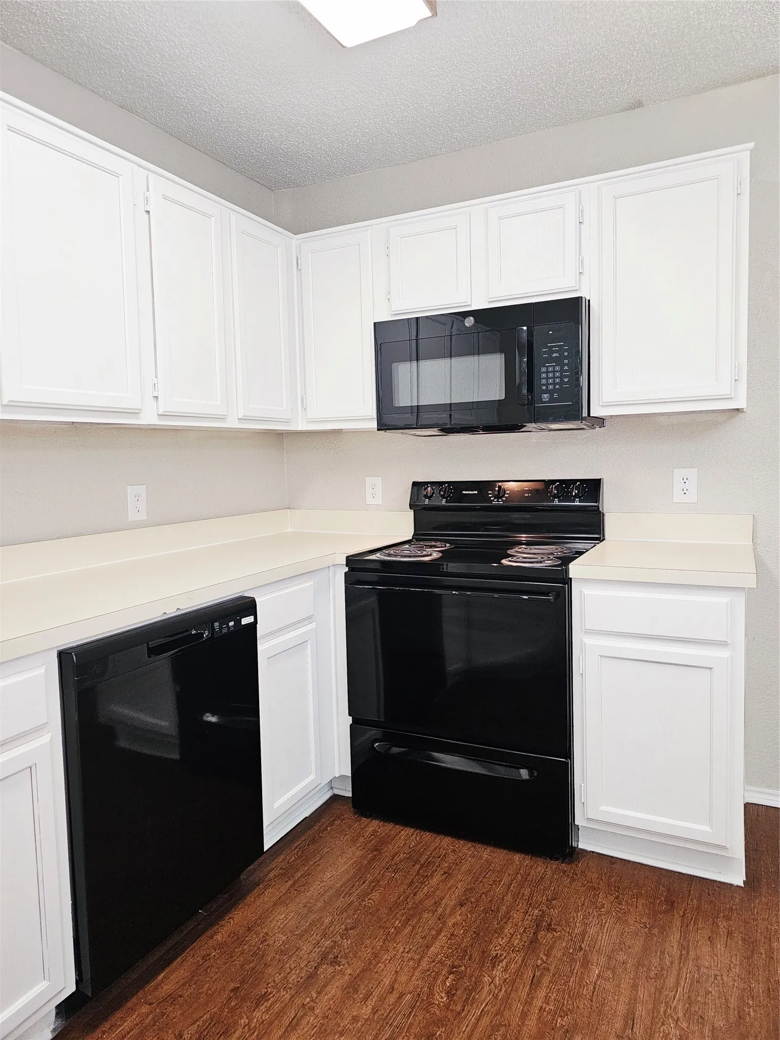 Corner kitchen featuring white upper and lower cabinetry, light-toned countertops, black appliances including a range and microwave, and wood-finish flooring