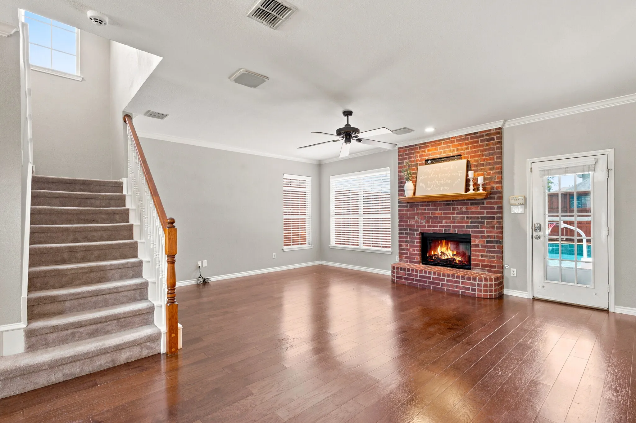Spacious living area featuring rich wood-finish flooring, a brick fireplace with a wood mantel, and a ceiling fan
