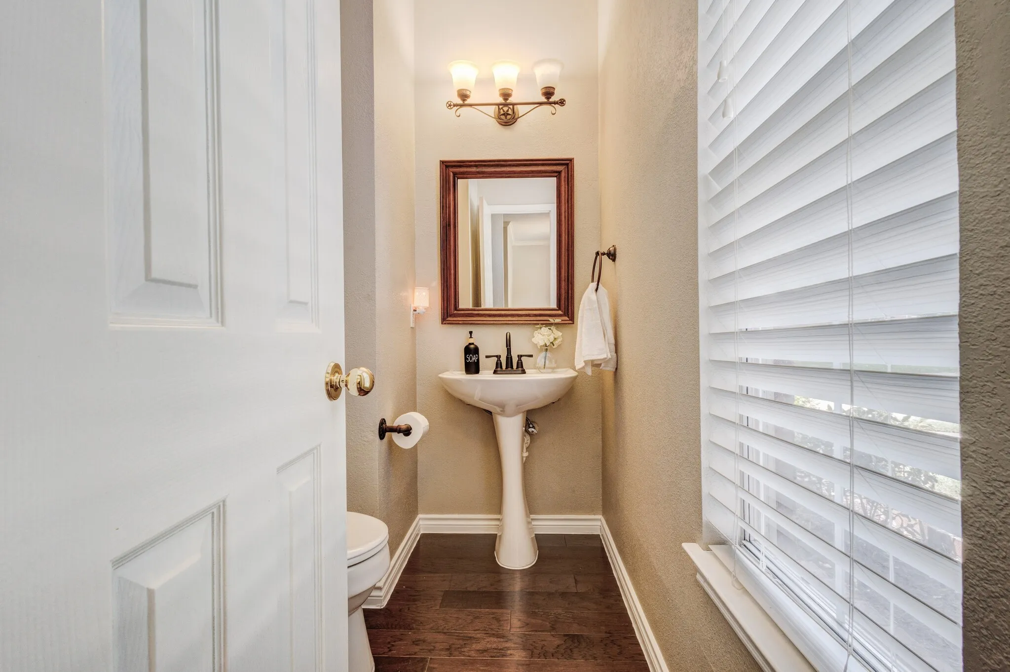 Pedestal sink with dark bronze faucet, wood-finish flooring, and a window with horizontal blinds