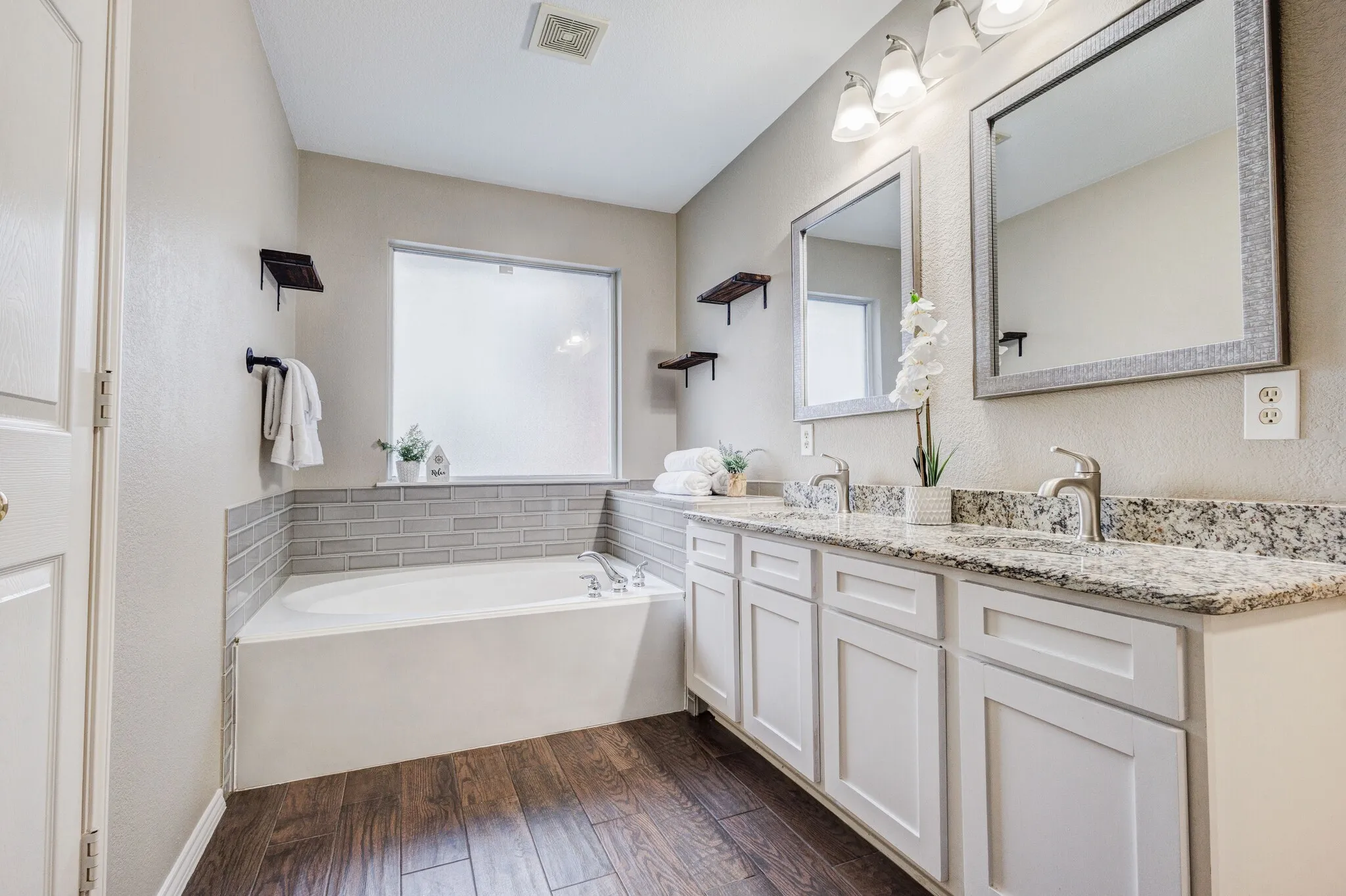 Bathroom with wood-finish flooring, a built-in tub featuring subway tile surround, a dual vanity with granite countertops, white cabinetry, and individual framed mirrors