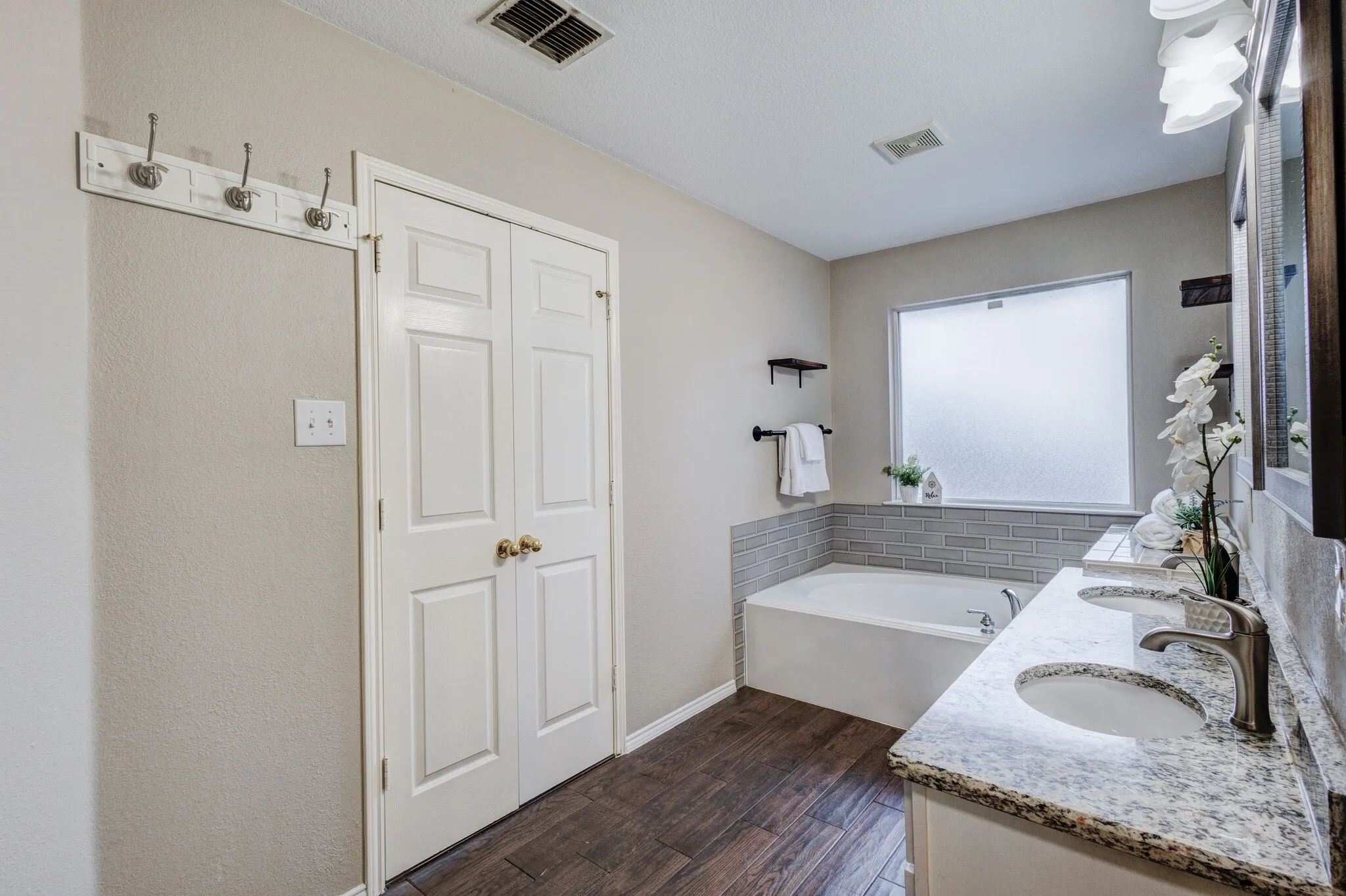 Bathroom featuring a double vanity with stone countertops, an integrated soaking tub with a tiled surround, wood-finish flooring, and a privacy window