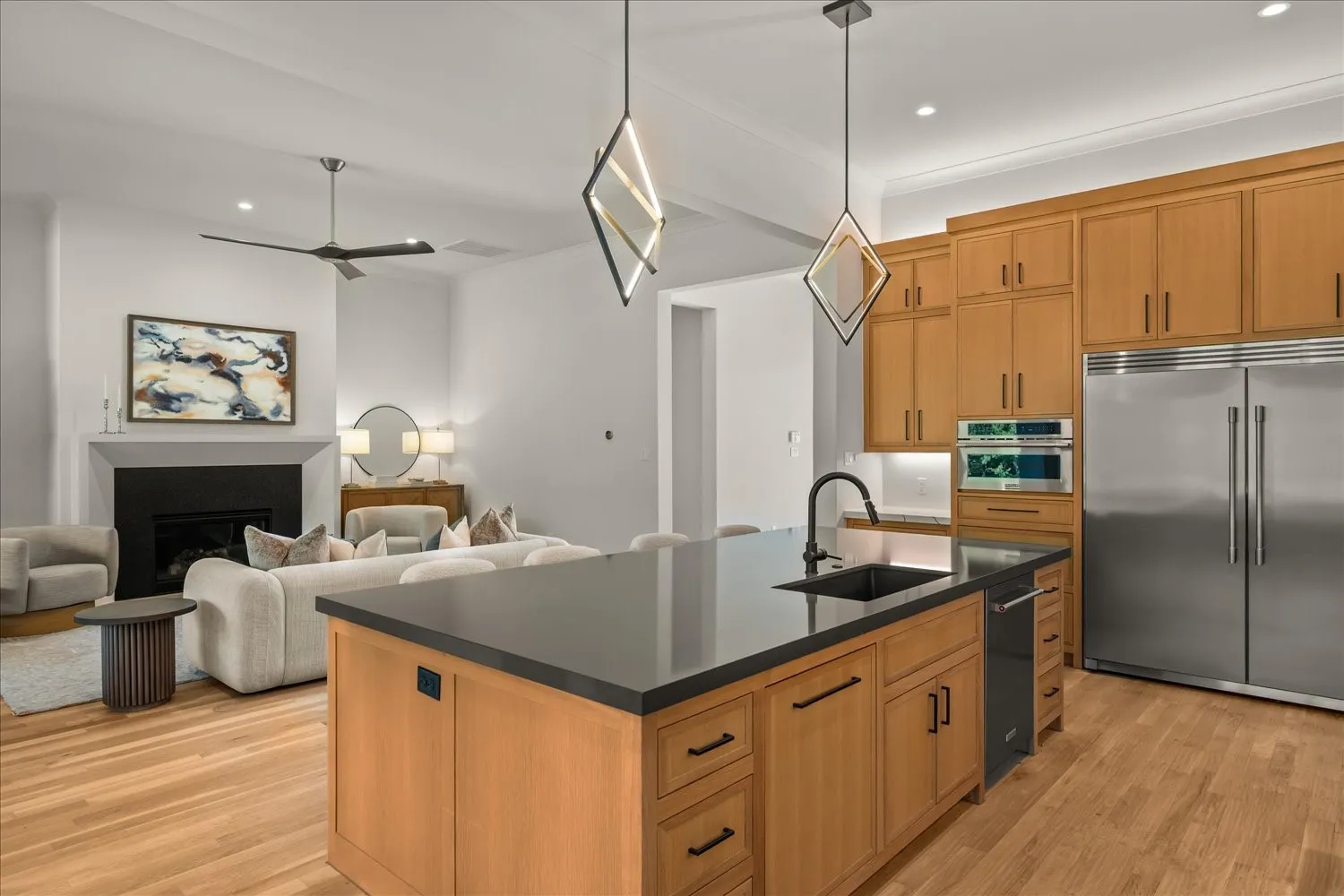 Open-concept living space featuring wood-finish flooring, a kitchen island with a dark countertop and undermount sink, light wood cabinetry, stainless steel appliances, and a living area with a fireplace