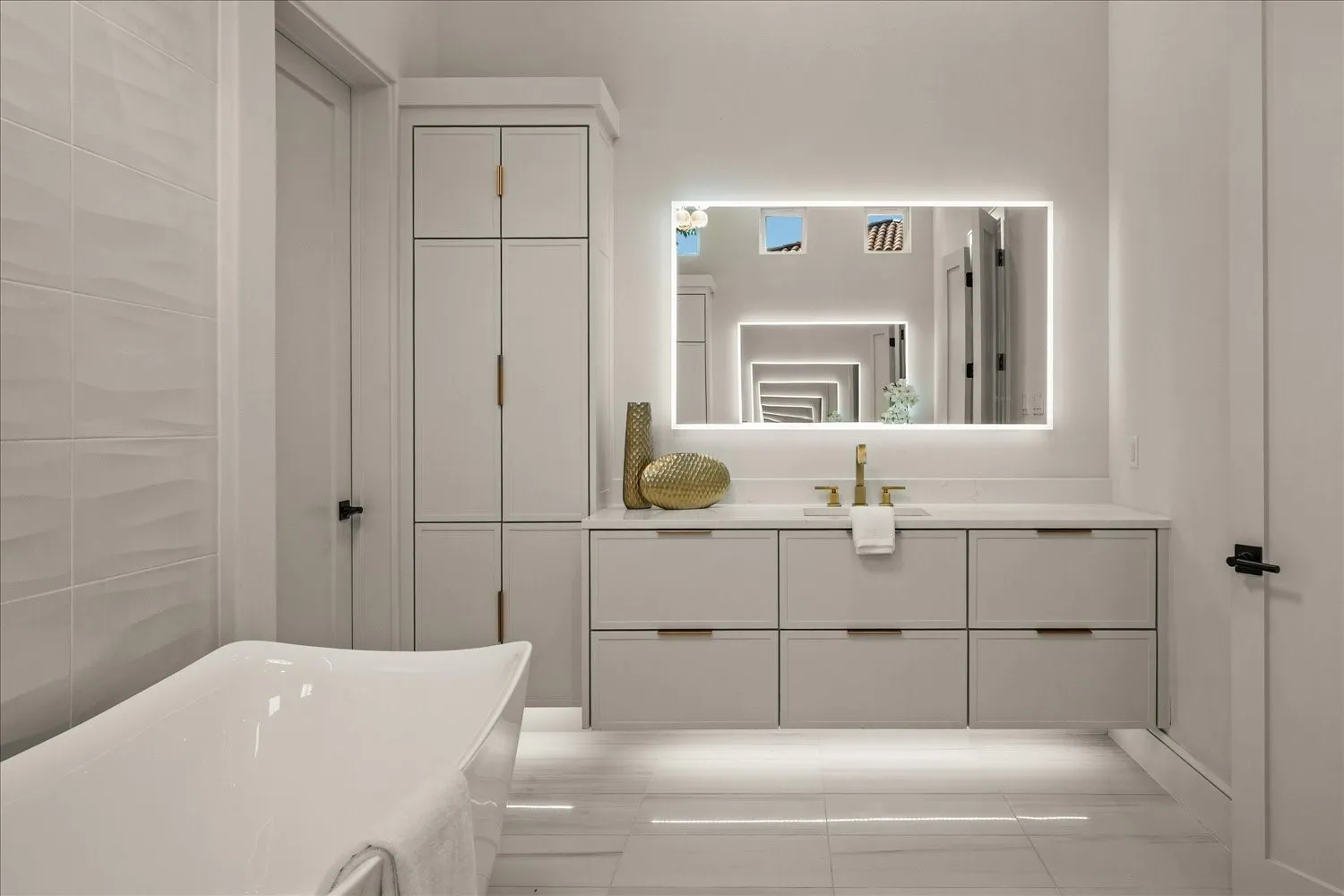 Freestanding soaking tub, sleek vanity with gold-tone hardware, illuminated mirror, built-in storage cabinetry, and large format floor tile
