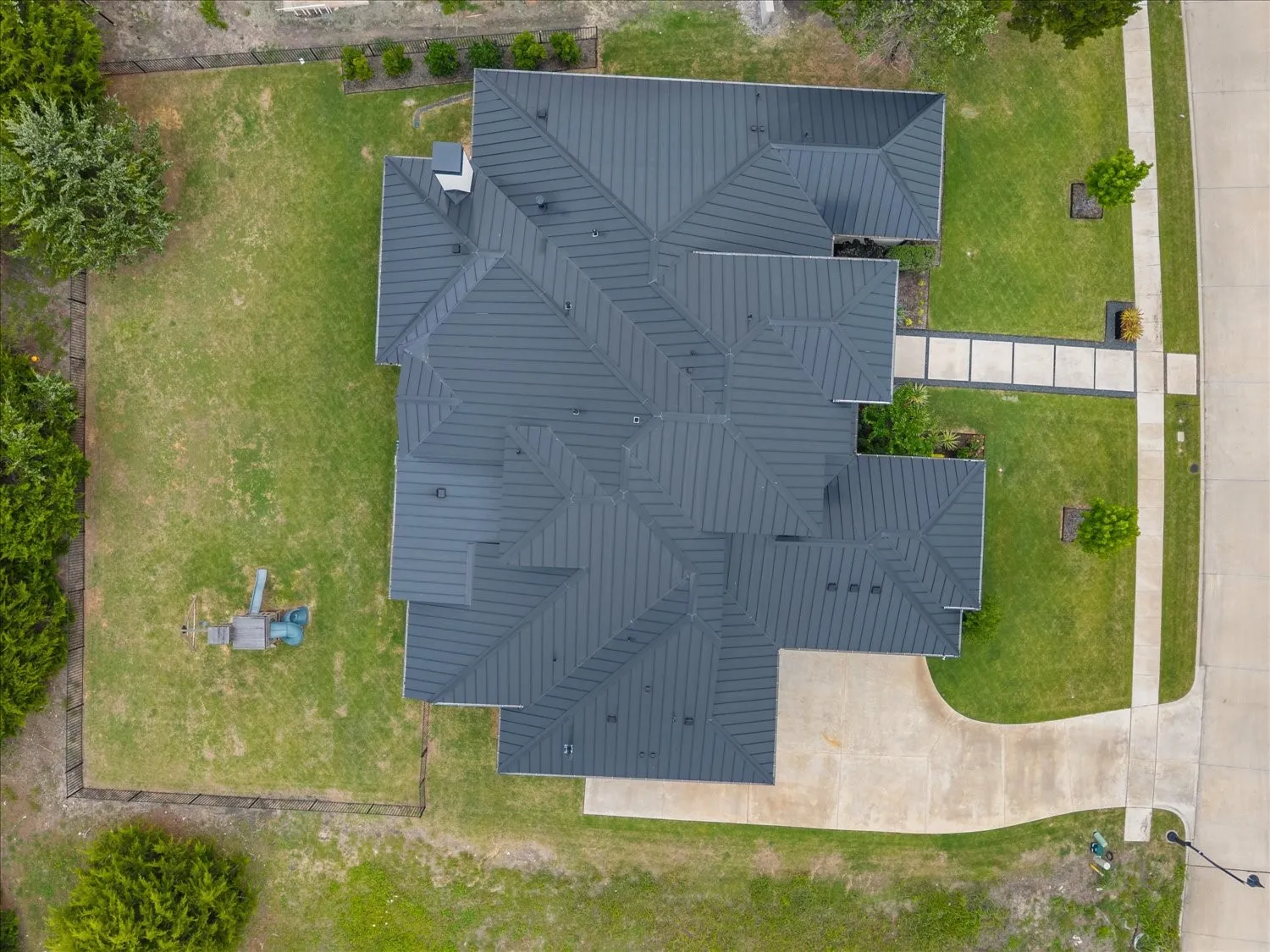 Multi-pitched dark gray roof, expansive green lawn, concrete driveway, concrete sidewalk, and a paver walkway