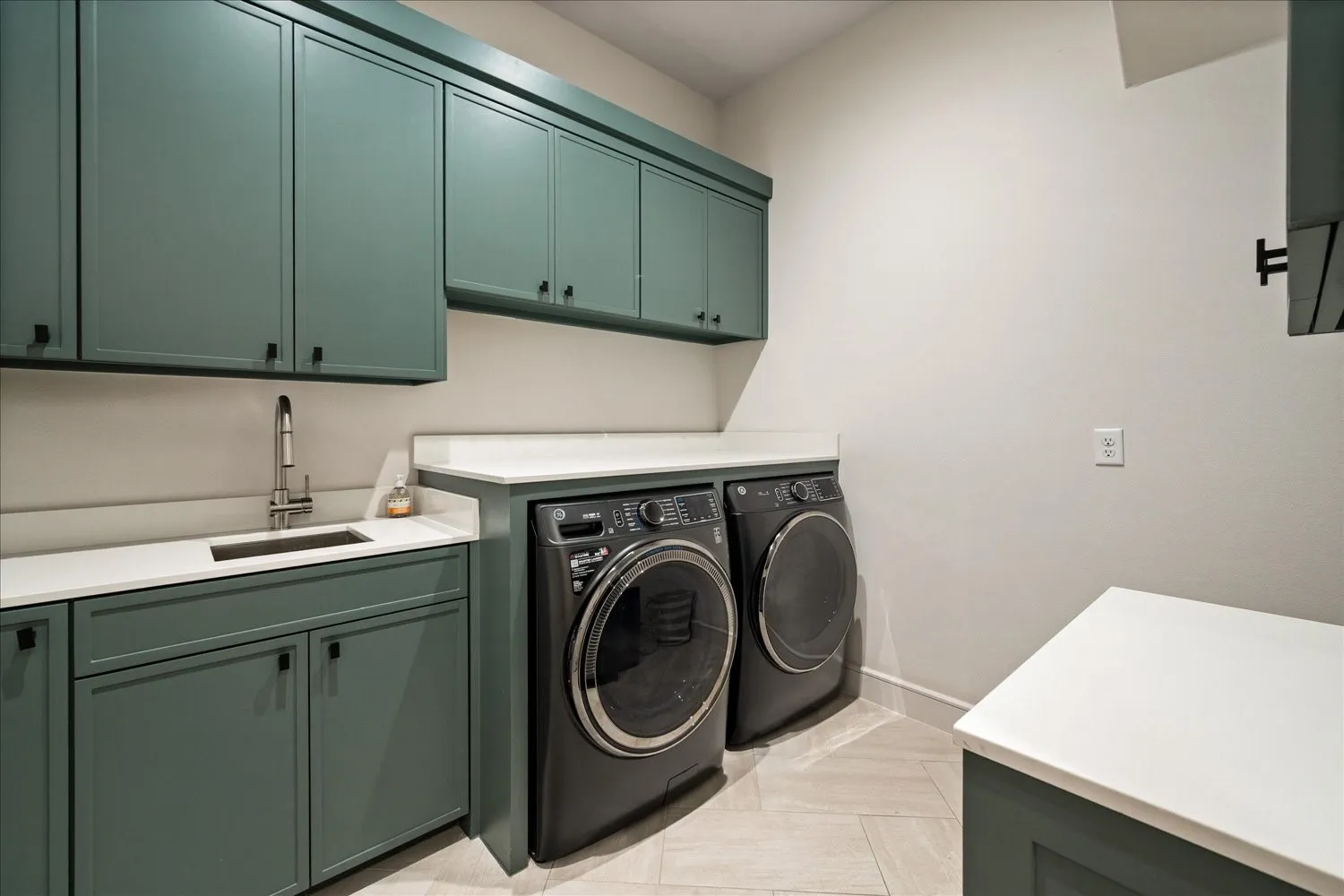 Laundry area featuring ample storage cabinetry in a muted green finish, white solid surface countertops, and an undermount sink with a gooseneck faucet