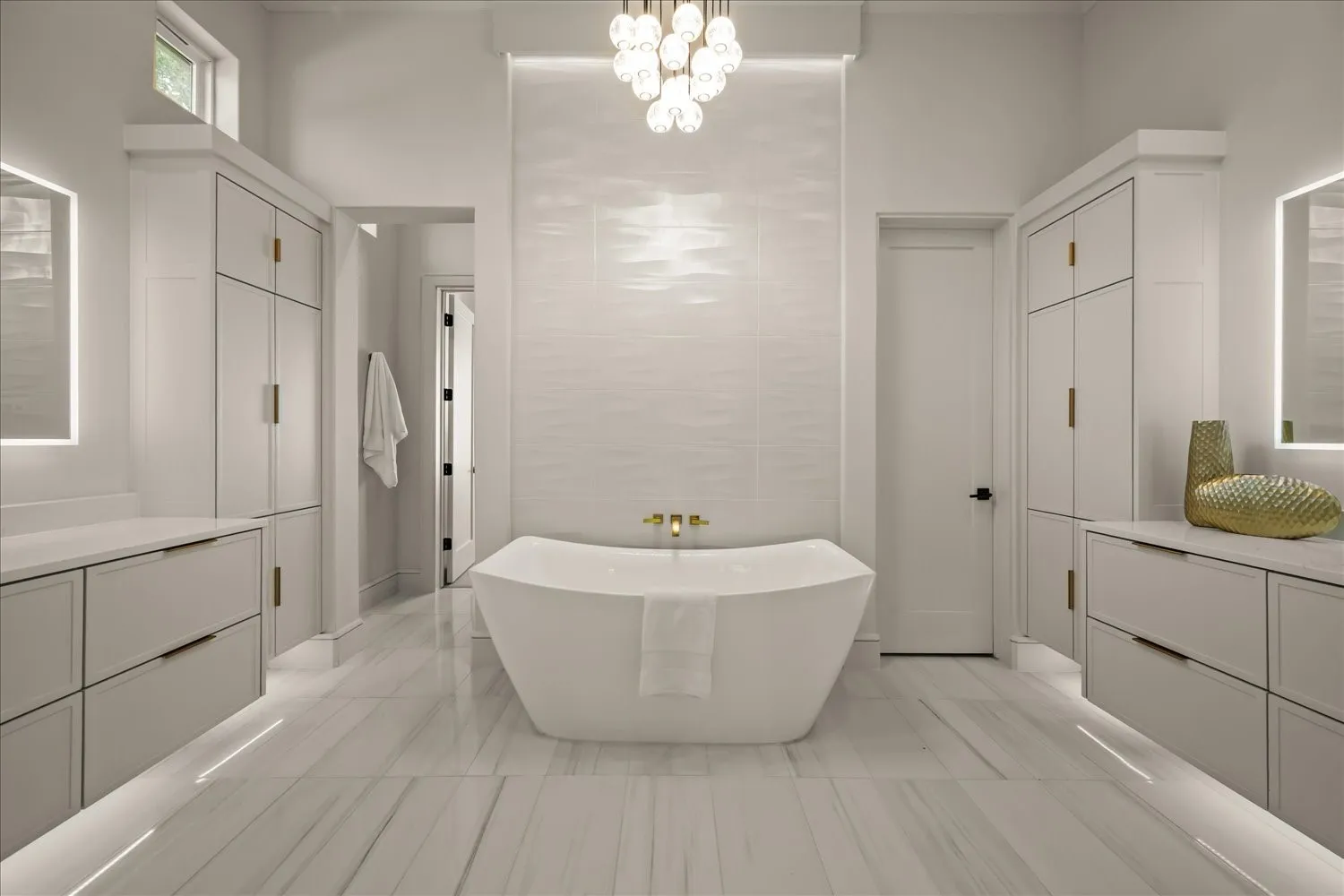 Freestanding soaking tub with a wall-mounted faucet, dual vanities with integrated lighting, tiled accent wall, and polished tile flooring
