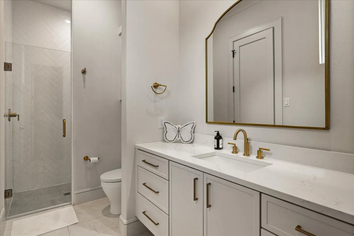 Bathroom featuring a frameless glass shower enclosure with chevron-patterned tile, a white vanity with integrated sink, and an arched mirror with brass-finish trim