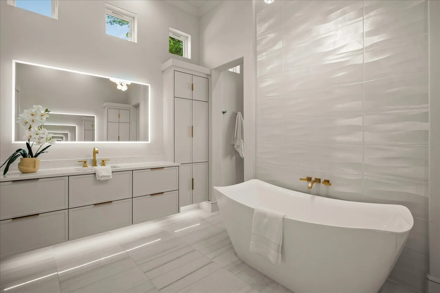Freestanding soaking tub, floor-to-ceiling textured wall tile, dual vanity with integrated sink, back-lit mirror, and built-in storage cabinetry