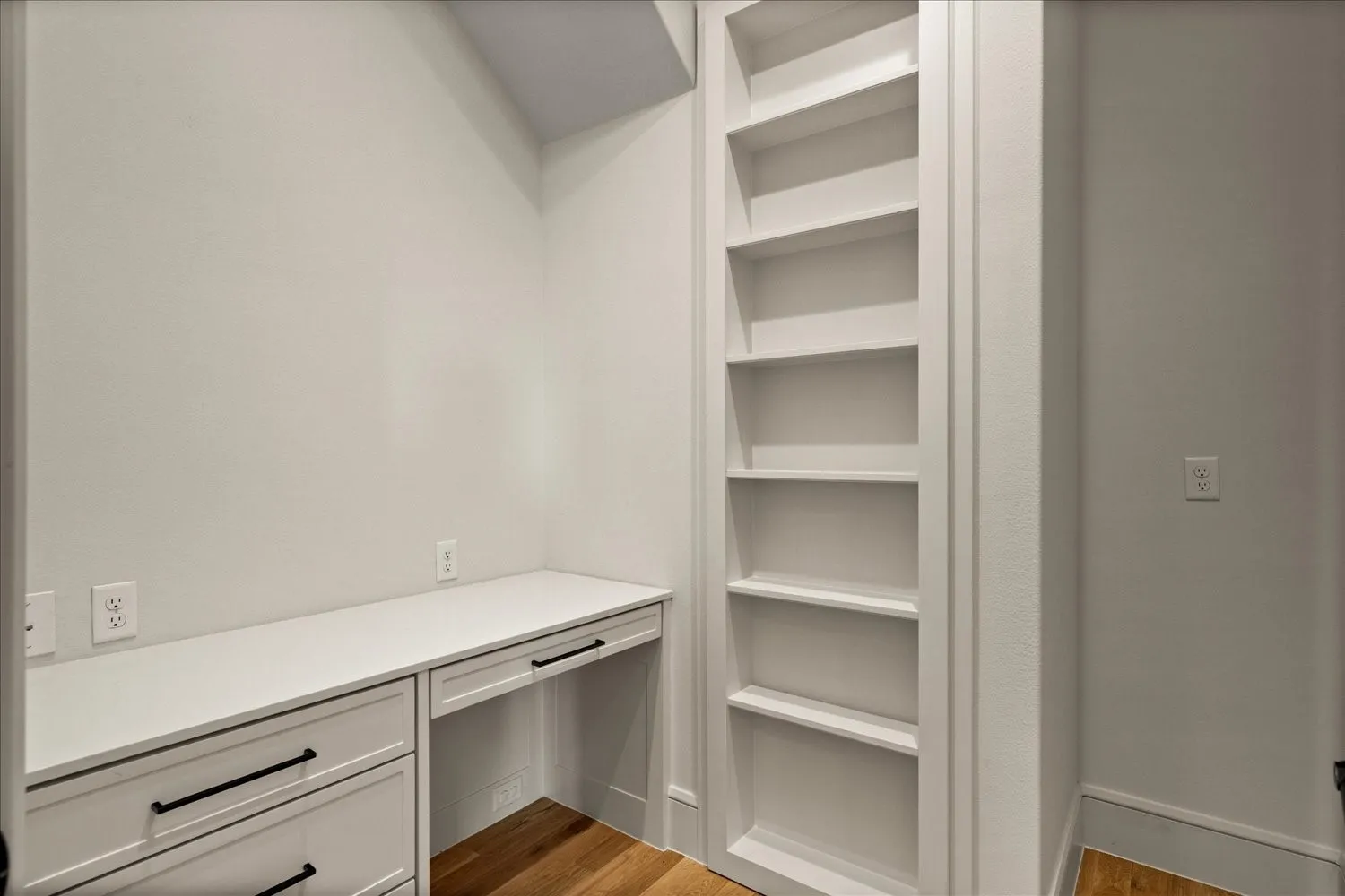 Built-in desk area featuring a white countertop and cabinetry with matte black hardware, integrated shelving, and wood-finish flooring