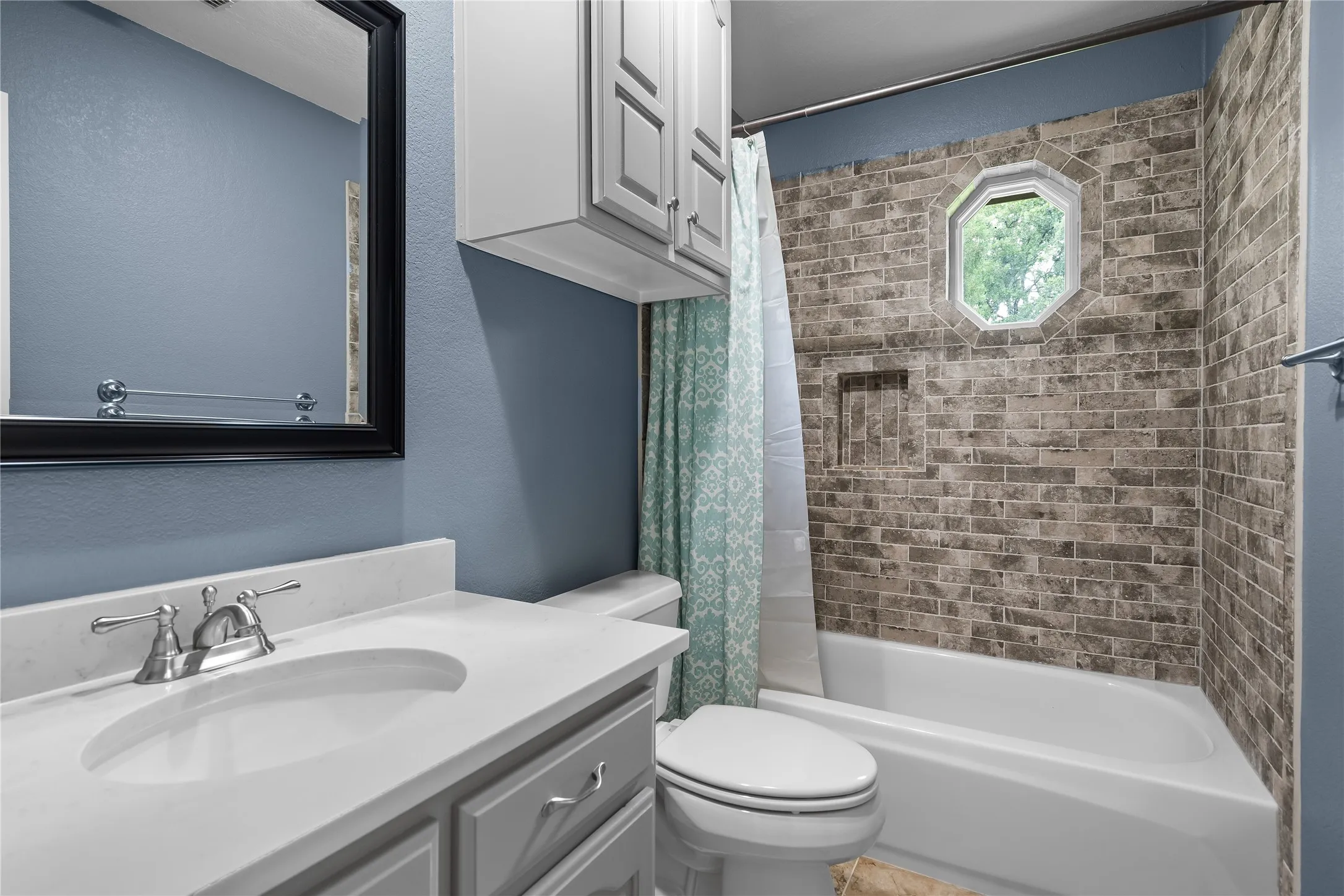 Bathroom featuring a white vanity with an integrated sink and chrome faucet, overhead cabinetry, a black-framed mirror, and a tiled shower surround with an octagonal window and built-in niche