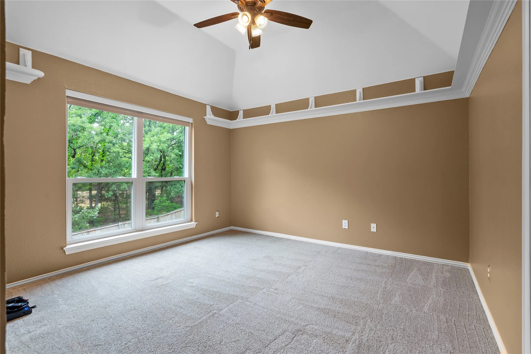 Room featuring a vaulted ceiling, ceiling fan with light fixture, decorative wall shelving, crown molding, and large window with exterior scenery