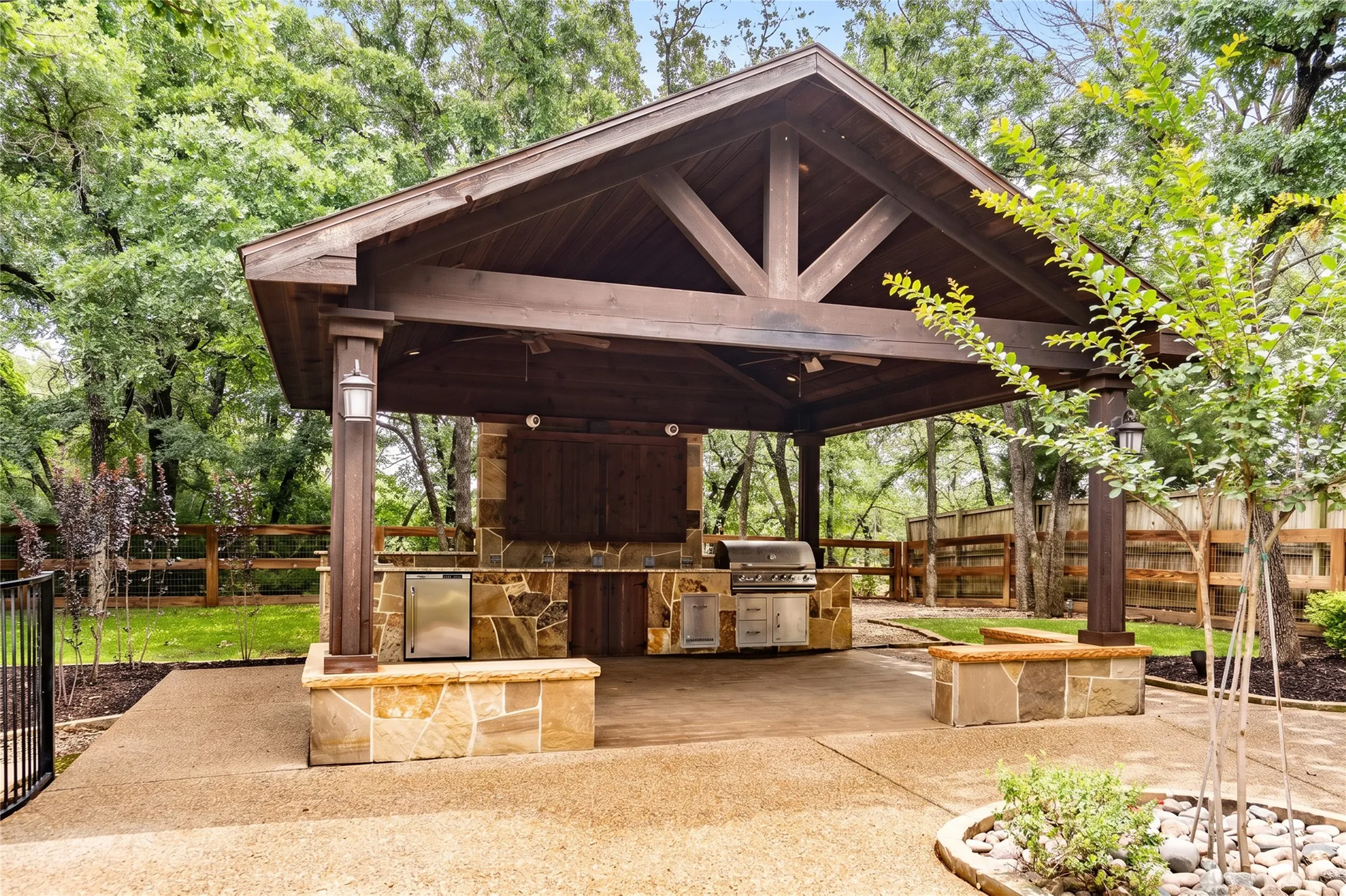 Expansive outdoor living area featuring a covered patio, exposed beam ceiling, stone-clad outdoor kitchen with built-in grill and refrigerator, and integrated lighting