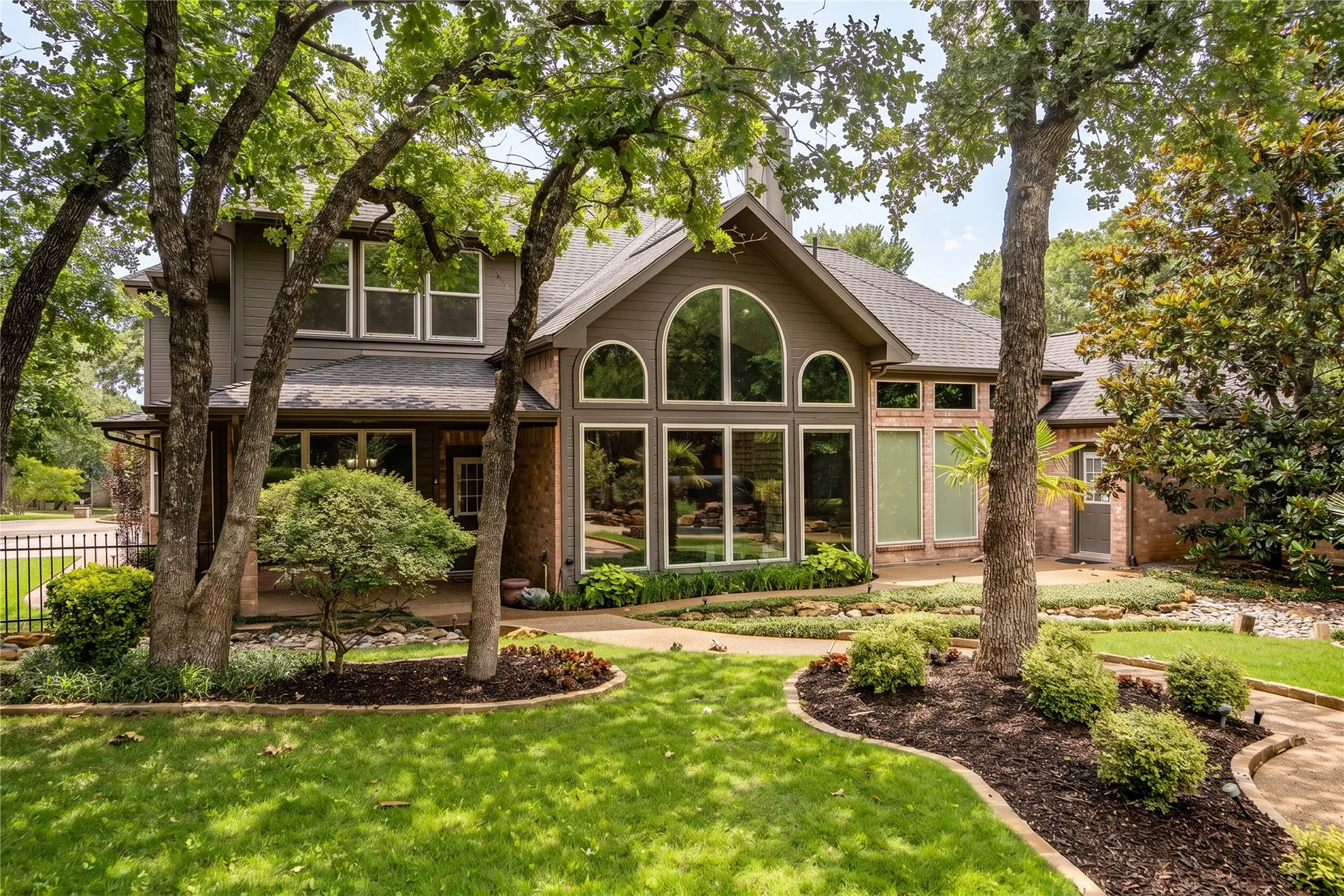 Exterior facade featuring brick and siding construction, a prominent arched window wall, multiple large windows, a dark shingle roof, and mature trees framing the property
