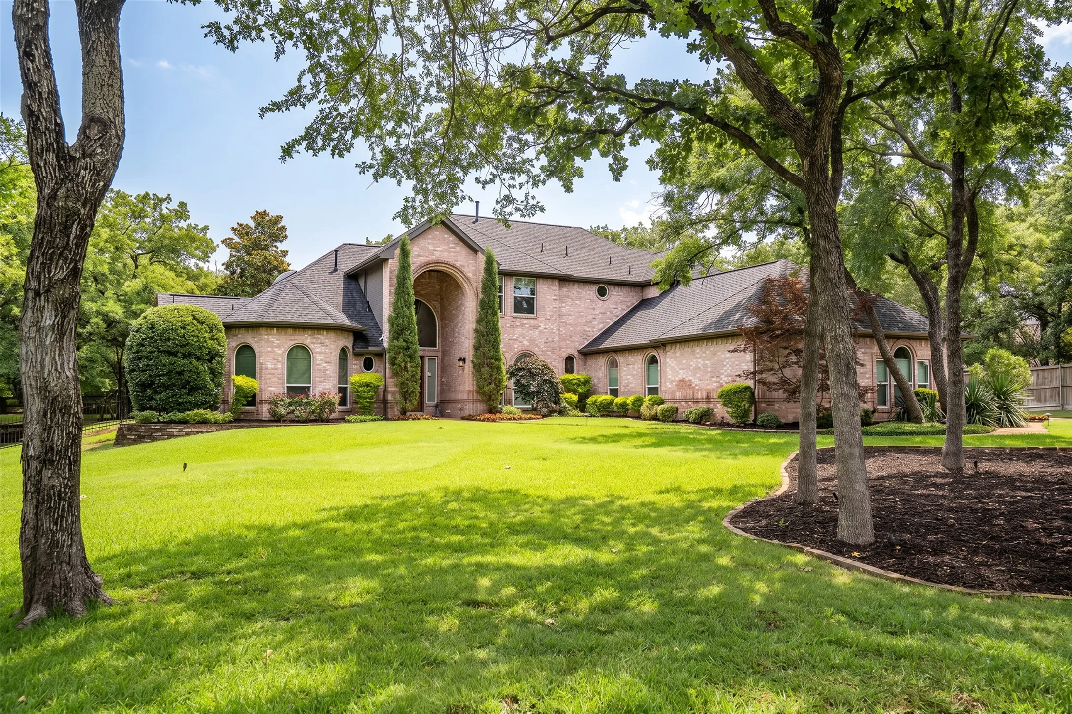 Traditional brick exterior with an arched entry, multiple arched windows, a dark shingled roof, a manicured lawn, and mature​​‌​​​​‌​​‌‌​‌‌​​​‌‌​‌​‌​‌​​​‌​​ trees