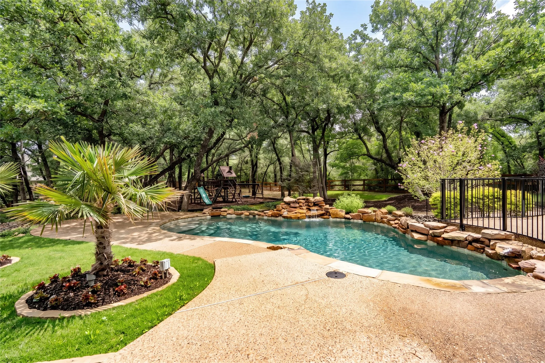 Curved swimming pool featuring a rock waterfall, surrounding stone hardscaping, and a wrought iron fence
