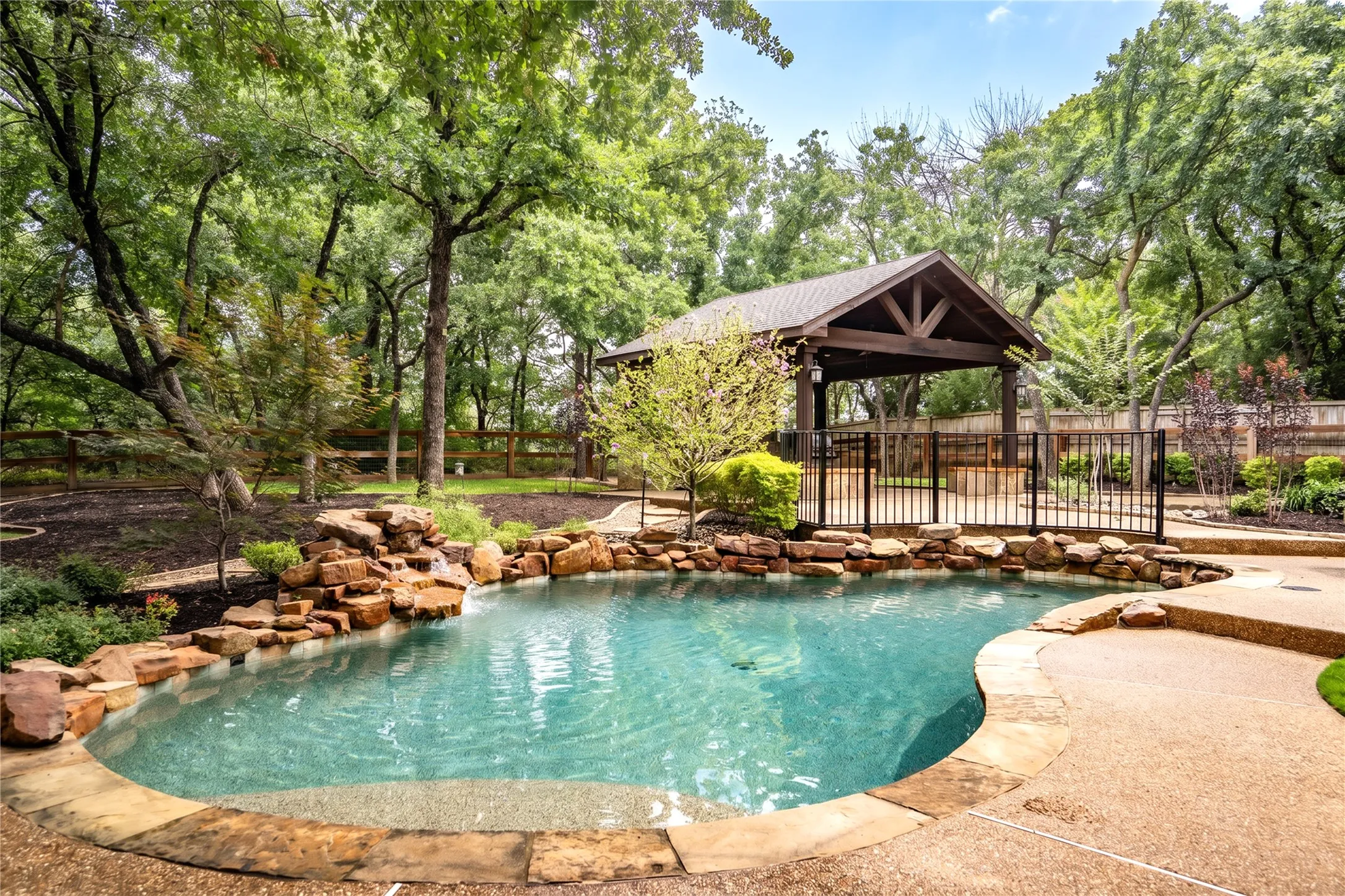 Expansive outdoor living area featuring a covered patio, exposed beam ceiling, stone-clad outdoor kitchen with built-in grill and refrigerator, and integrated lighting