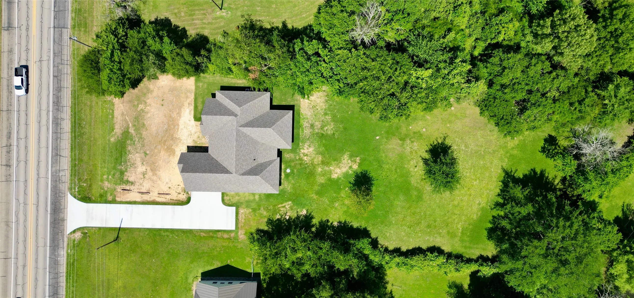 Newly constructed residence with a complex roofline, a newly poured concrete driveway, and surrounding green space with mature trees