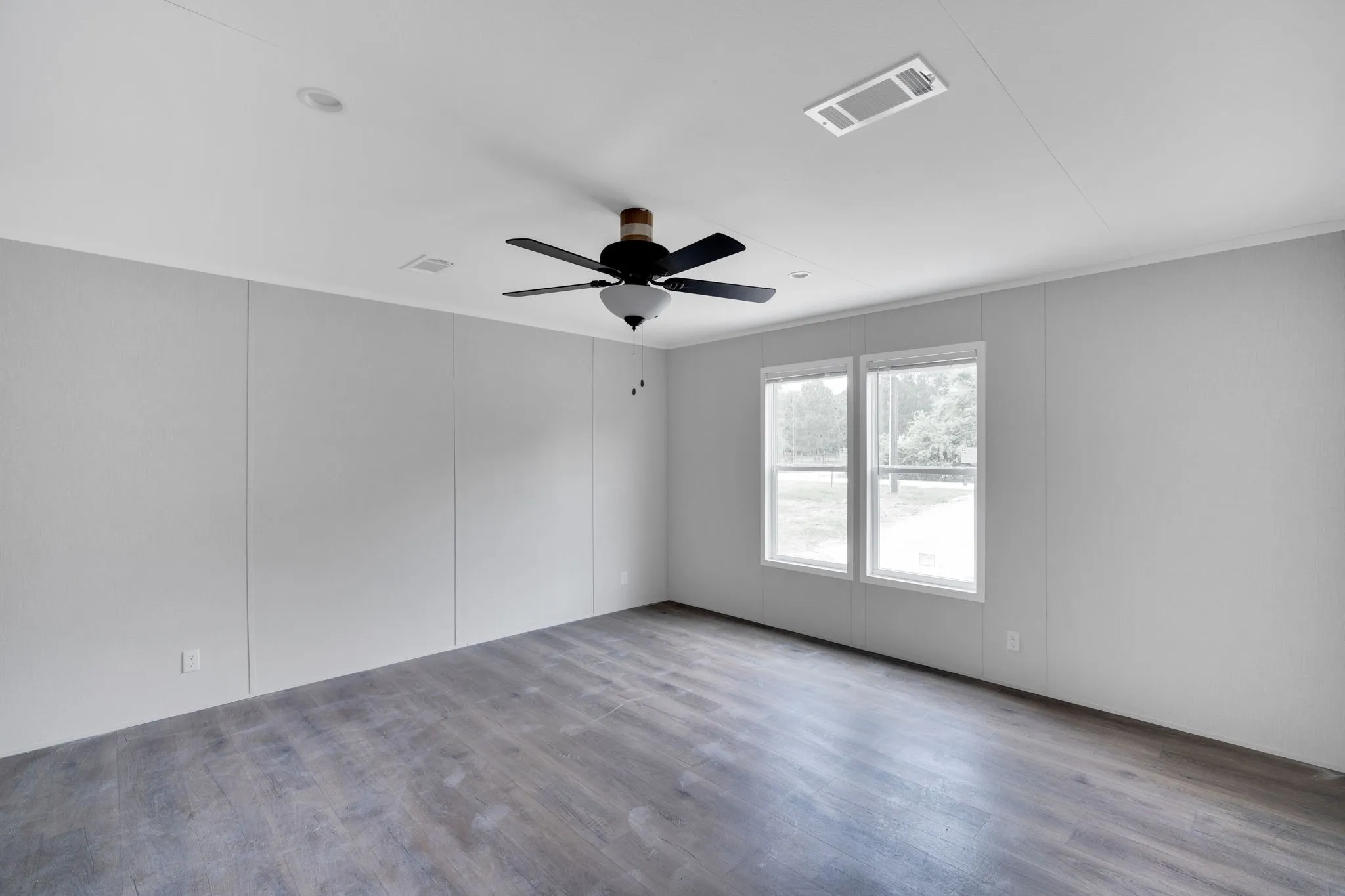 Spacious room featuring wood-finish flooring, light grey paneled walls, and a ceiling fan with integrated lighting