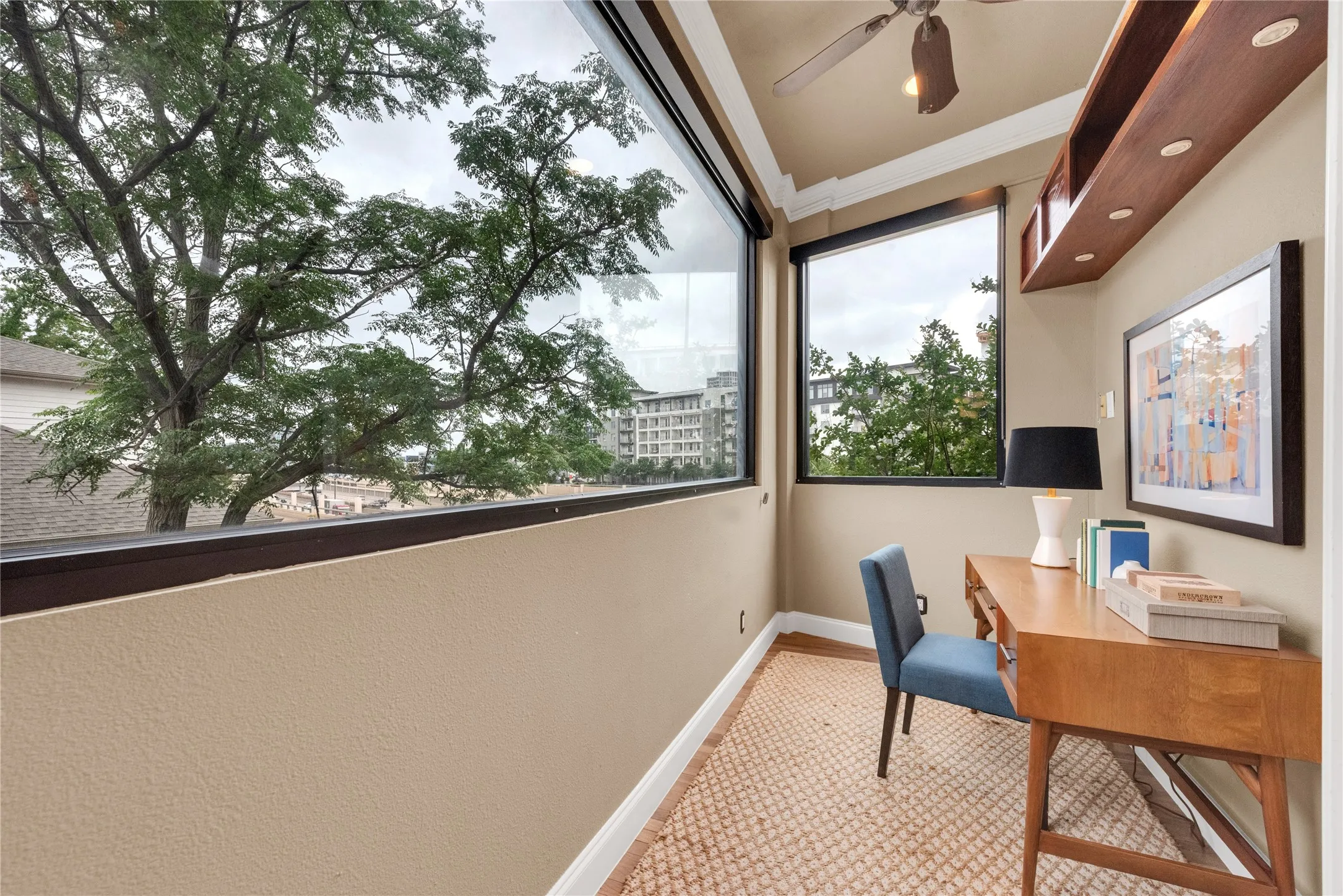 Dedicated office space featuring large windows, wood-finish flooring, built-in shelving with integrated lighting, a ceiling fan, and crown molding