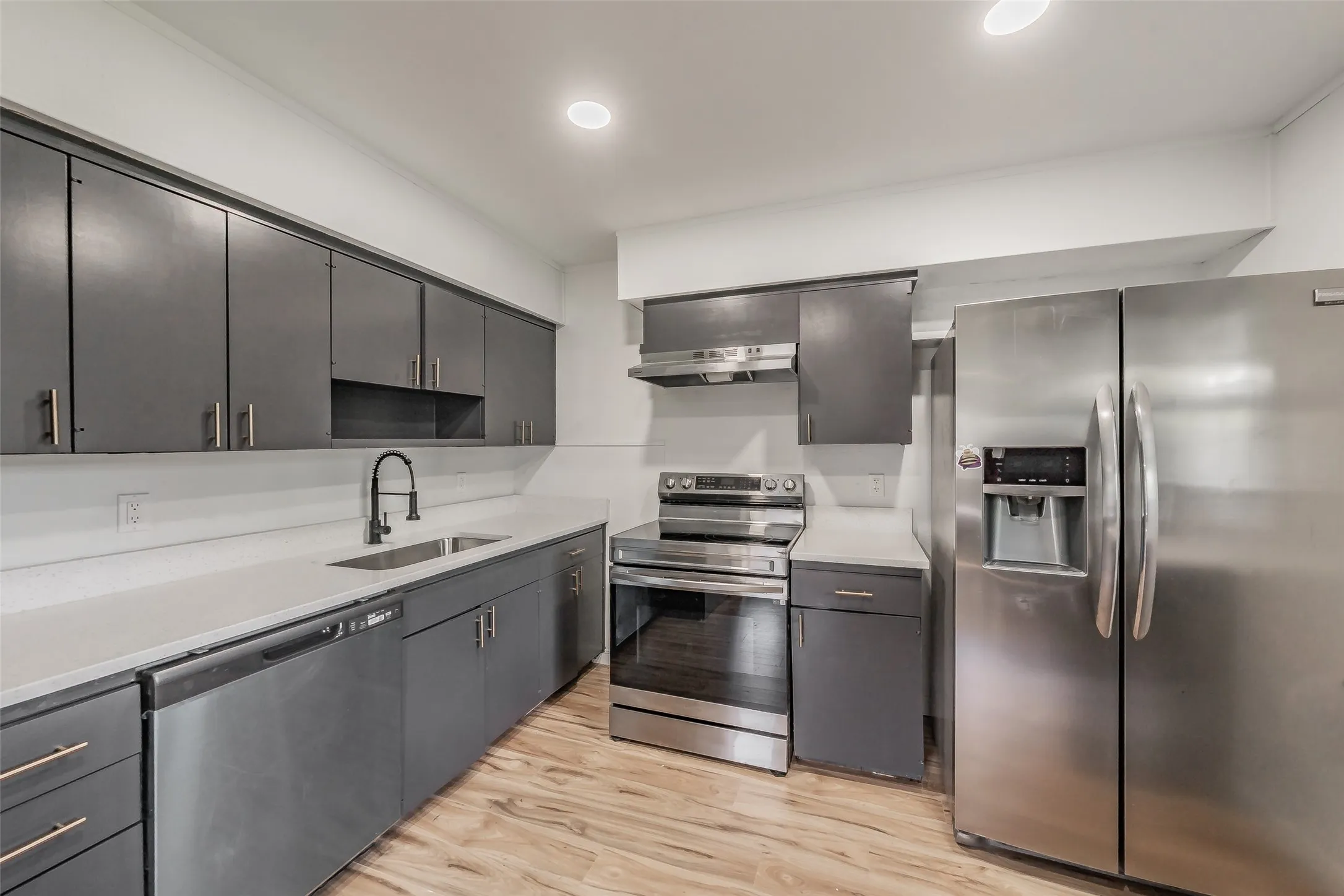 Modern kitchen featuring stainless steel appliances, grey cabinetry with gold-tone hardware, white countertops, a matte black faucet, and wood-finish flooring