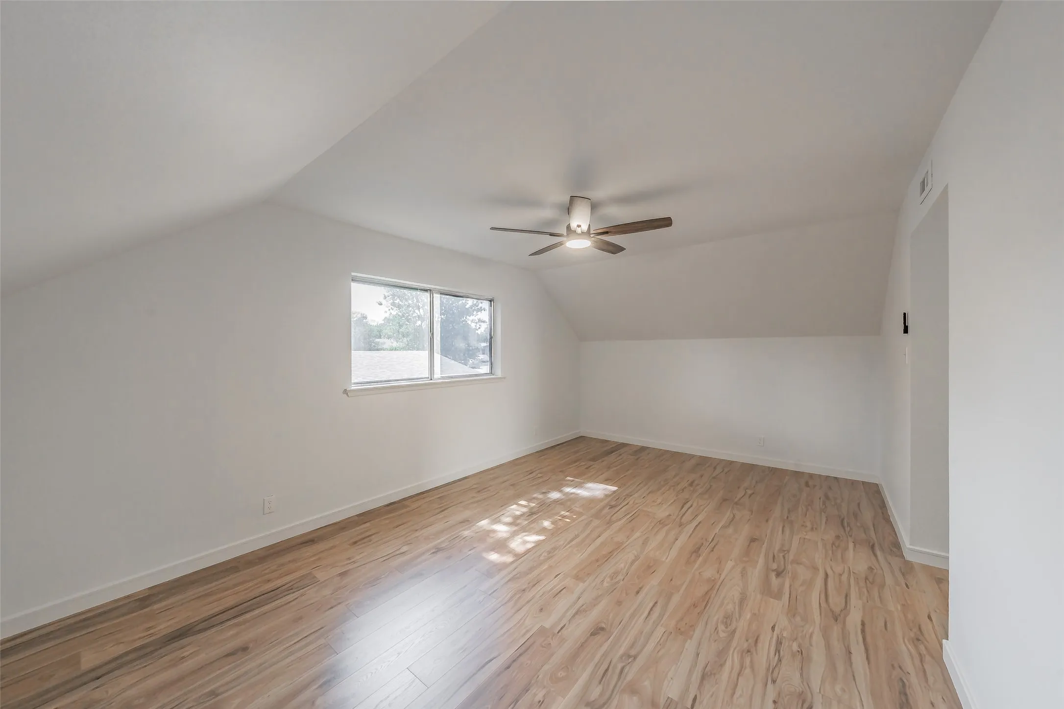 Spacious room with sloped ceilings, wood-finish flooring, a ceiling fan, and a large window providing ample natural light