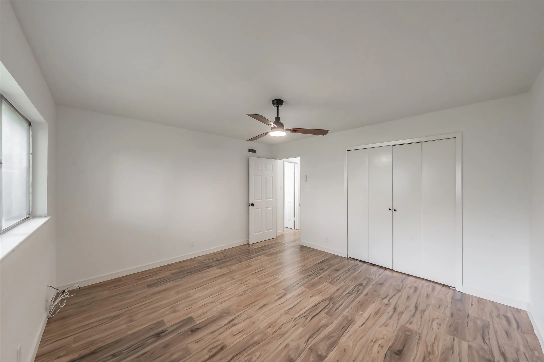 Room featuring wood-finish flooring, a ceiling fan with integrated lighting, a window with a roller shade, and bi-fold closet doors