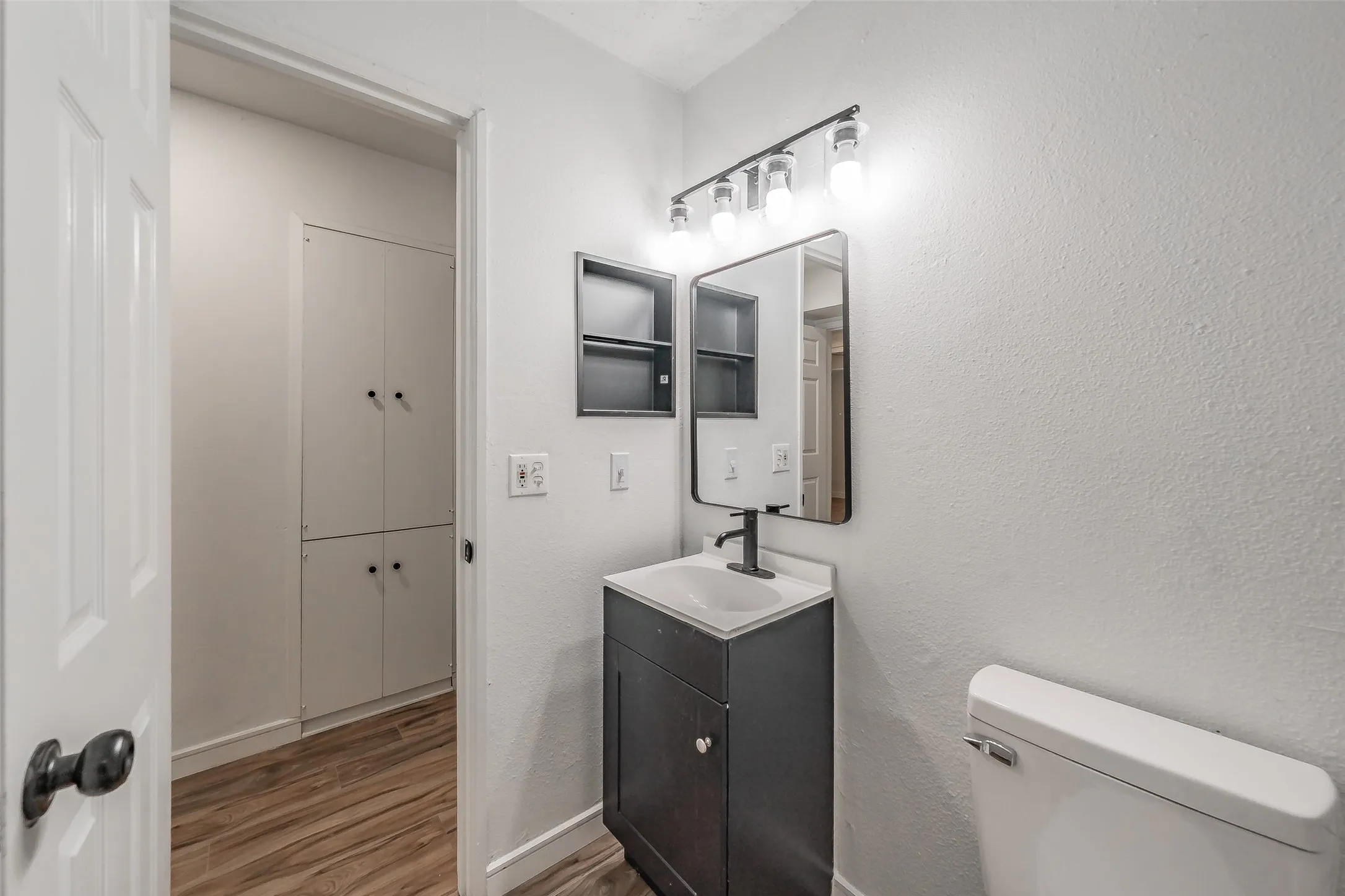 Bathroom featuring wood-finish flooring, a dark vanity with white countertop, a black-framed mirror, and an overhead light fixture