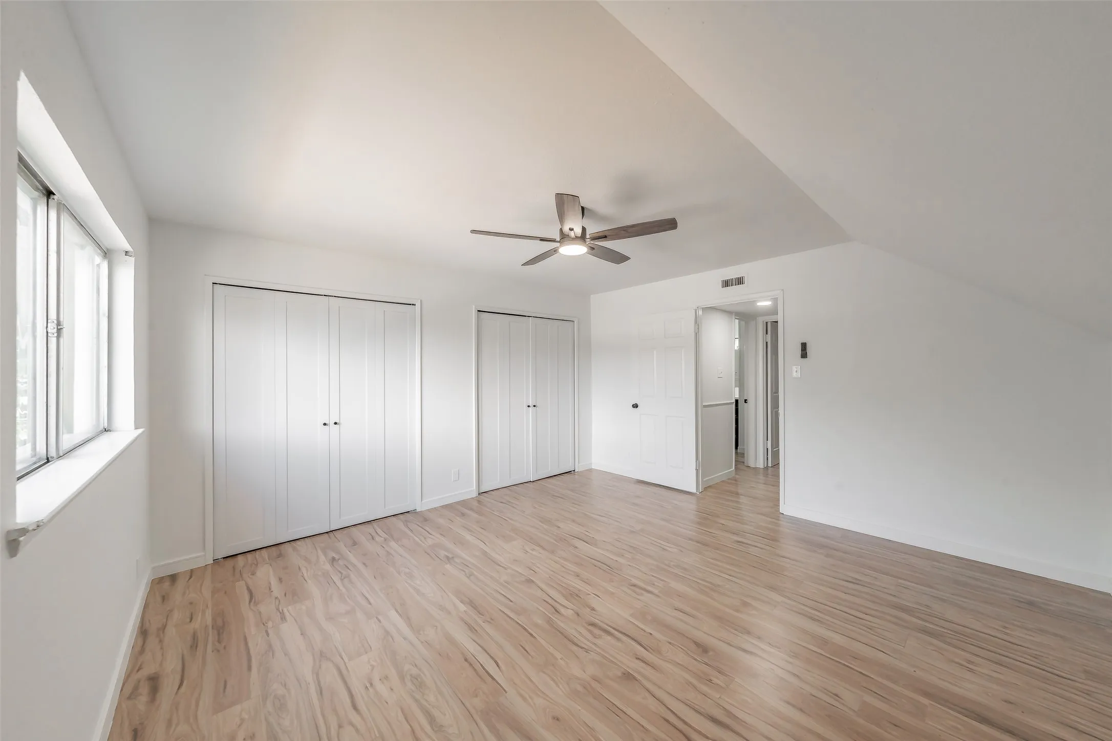 Bright room featuring wood-finish flooring, a ceiling fan with integrated lighting, multiple bi-fold closet doors, and a window with white trim