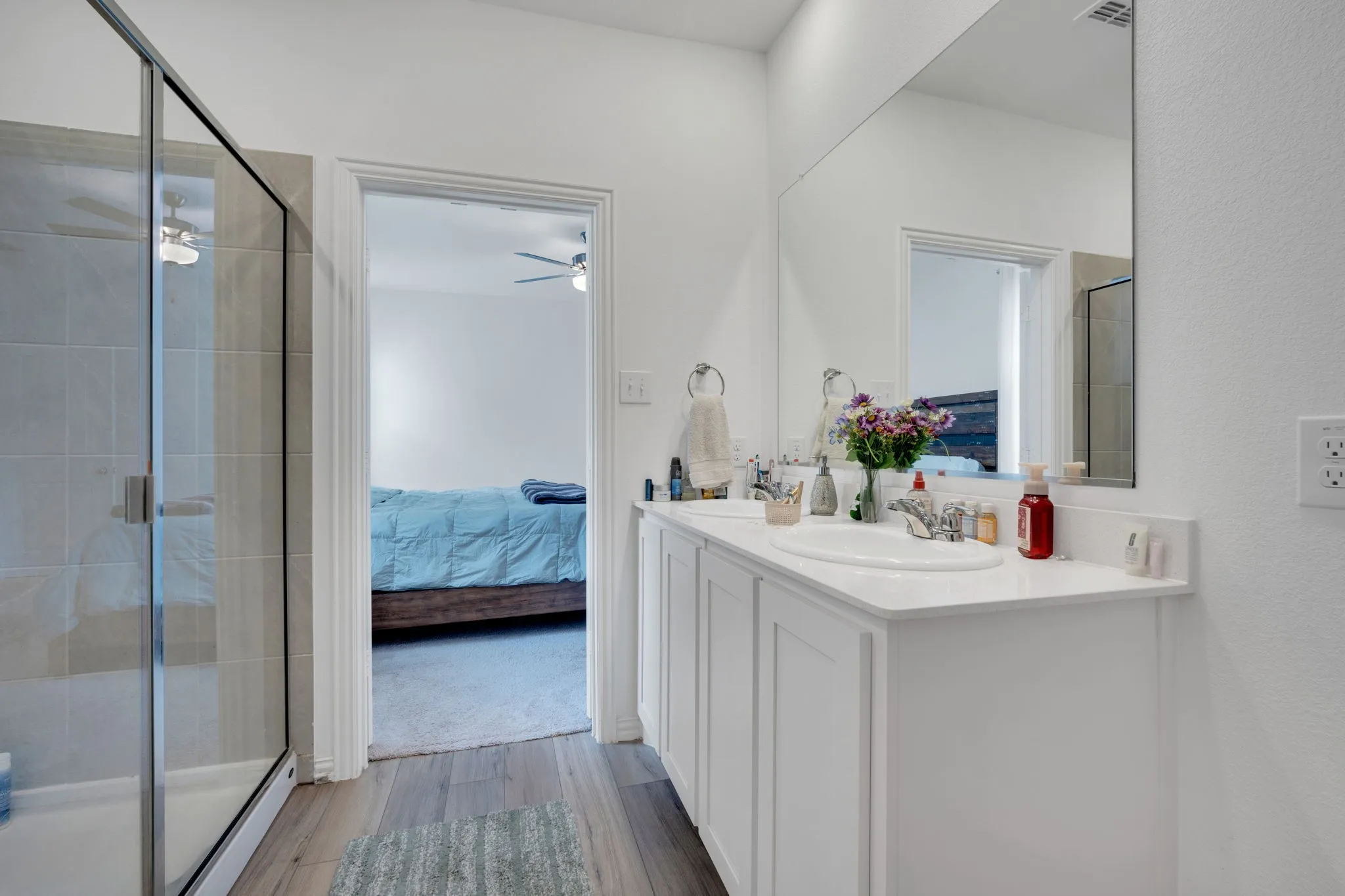 Bathroom featuring a double vanity with white cabinetry and an integrated countertop, a large wall-mounted mirror, a glass-enclosed shower with tiled walls, and wood-finish flooring