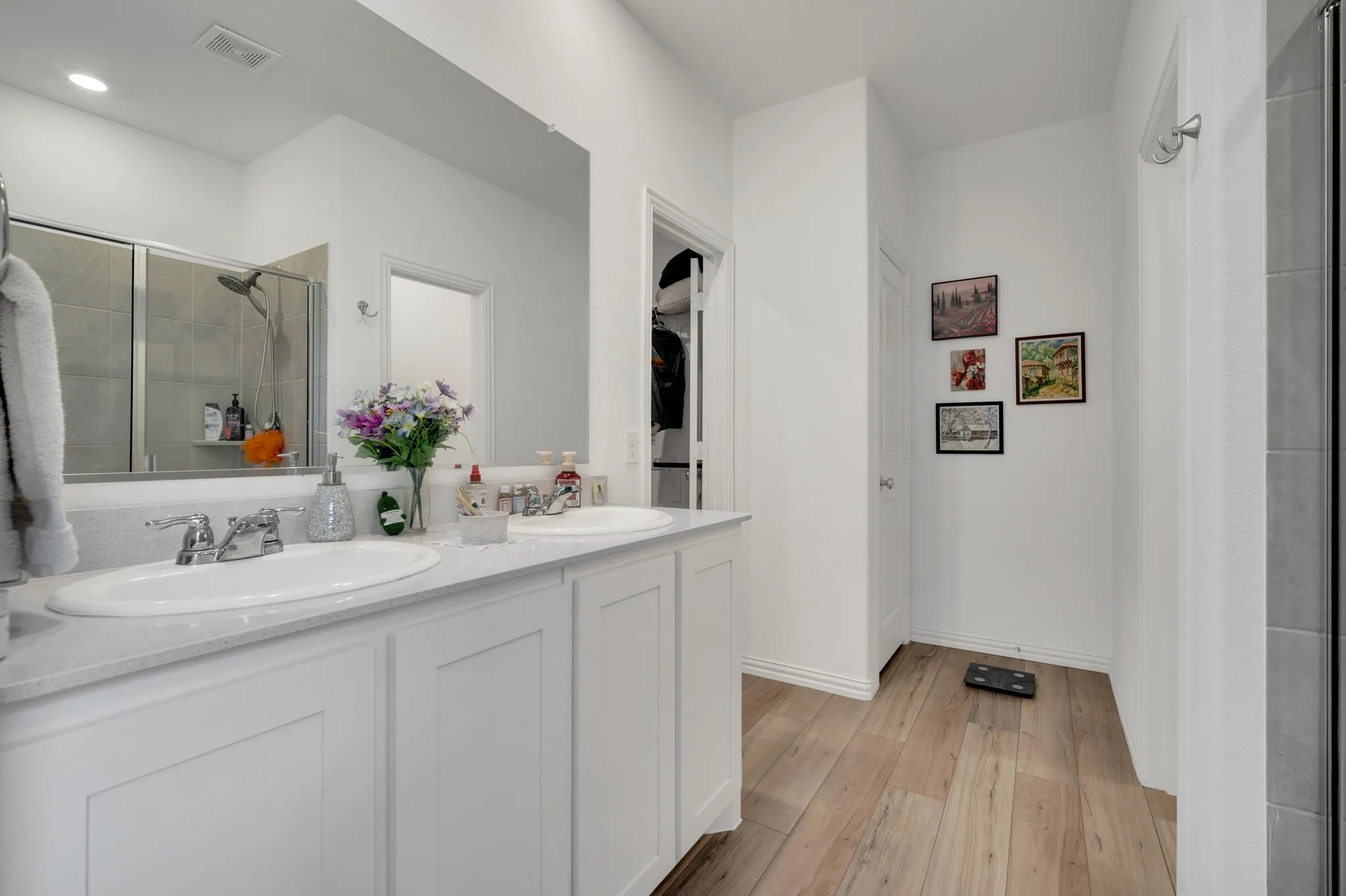 Bathroom featuring a double vanity with white cabinetry and an integrated countertop, dual undermount sinks with chrome fixtures, wood-finish flooring, and an enclosed shower with a glass door