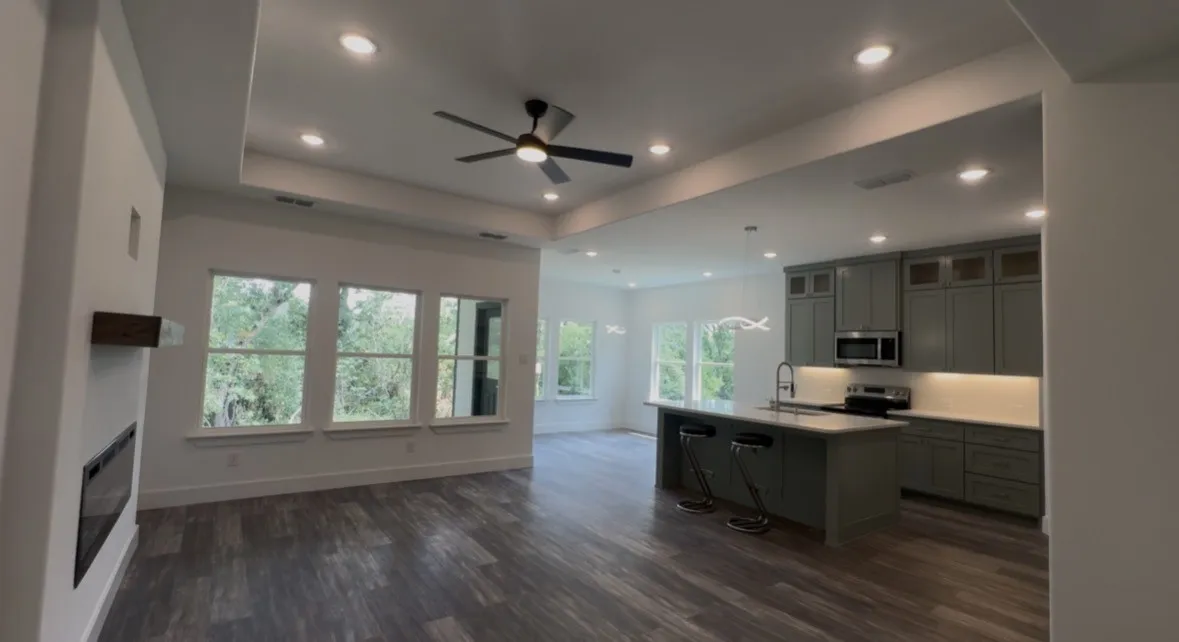 Open-concept living area featuring wood-finish flooring, a contemporary fireplace with a wood mantle, and a tray ceiling with recessed lighting and a ceiling fan