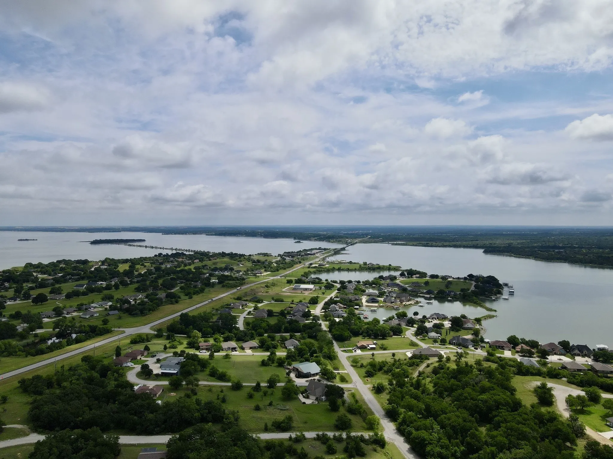 Expansive aerial perspective showcasing a community situated along a large body of water, featuring a peninsula with a connecting road, numerous properties with substantial green spaces, and dense tree coverage