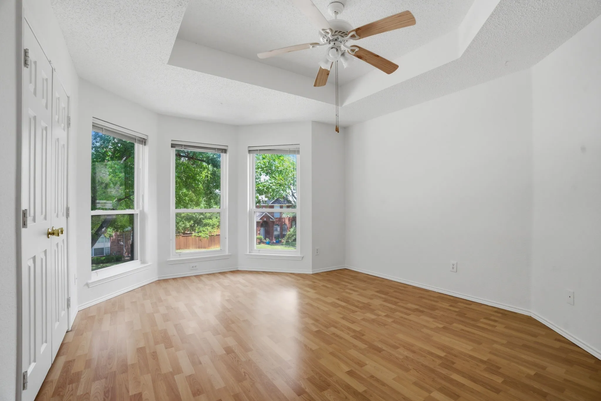 Light-filled room featuring a bay window with three vertical windows, wood-finish flooring, a tray ceiling, and a ceiling fan