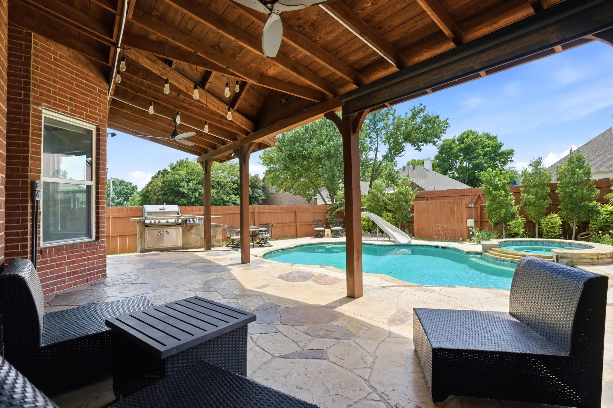 Covered patio featuring a wood-beam ceiling with string lighting, brick exterior wall, a built-in outdoor kitchen with grill, a freeform swimming pool with slide, and an integrated spa