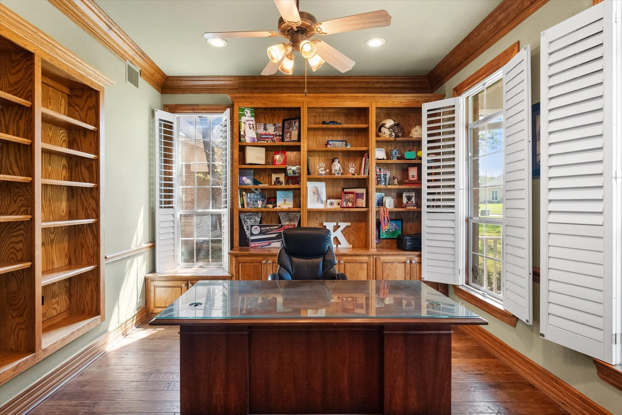 Beautifully appointed **home office with rich wood flooring** and **custom built-in cabinetry**, perfect for working from home. This light-filled space features **walls of windows with plantation shutters**, allowing for abundant natural light while maintaining privacy. The **extensive built-ins and shelving** provide ideal storage and display options, combining functionality with timeless style. A perfect blend of **executive office, study, or flex space**, designed for productivity and comfort in today’s modern lifestyle.
