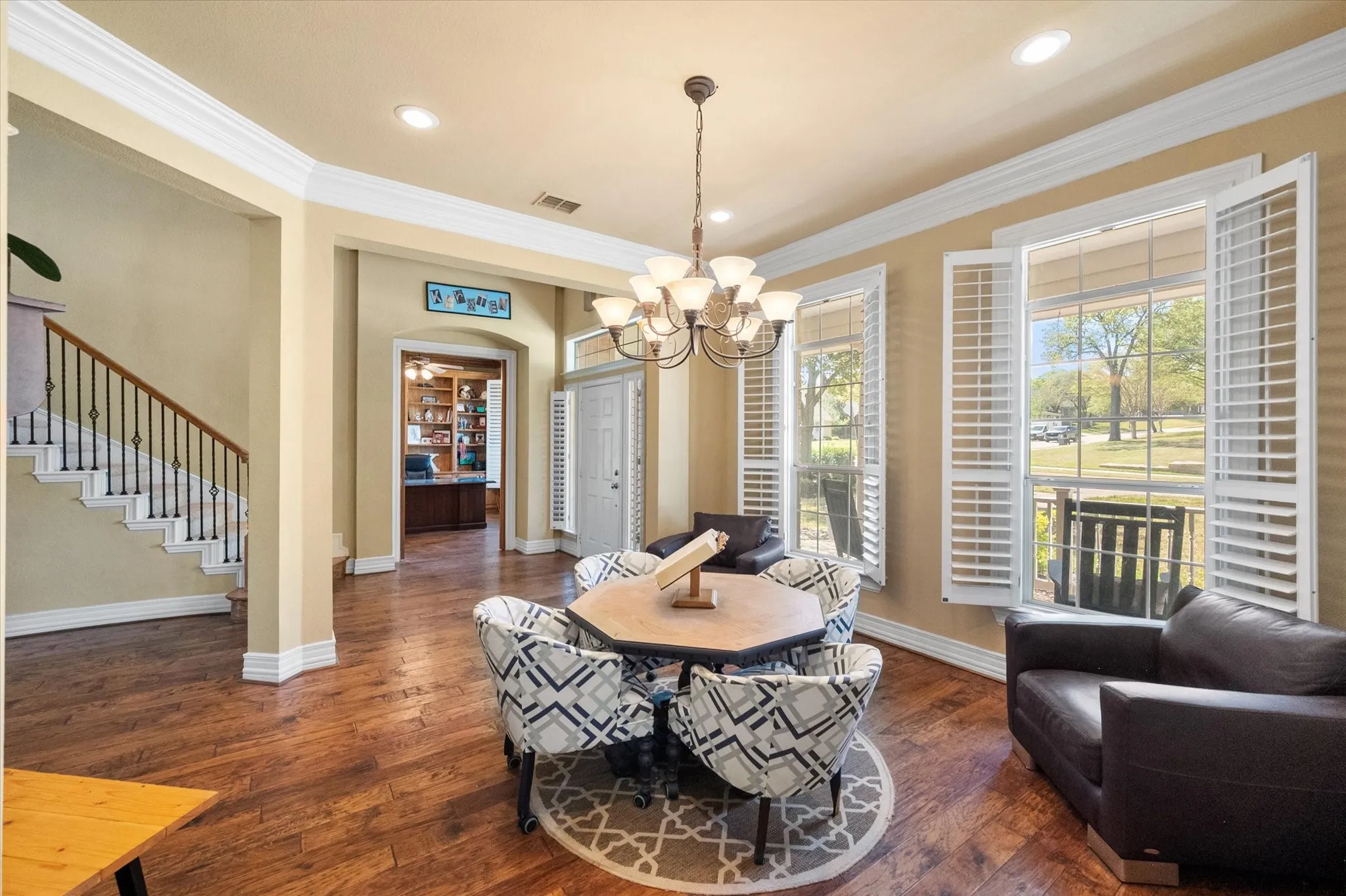 Stunning **formal dining room with open-concept design** seamlessly flows into the grand entry, showcasing a **sweeping staircase with wrought iron spindles** for a dramatic and elegant backdrop. Featuring **rich wood flooring, crown molding, and a wall of windows with plantation shutters**, this light-filled space is perfect for hosting gatherings and entertaining guests. The open layout enhances connectivity while maintaining defined spaces, creating an ideal setting for both everyday dining and special occasions.