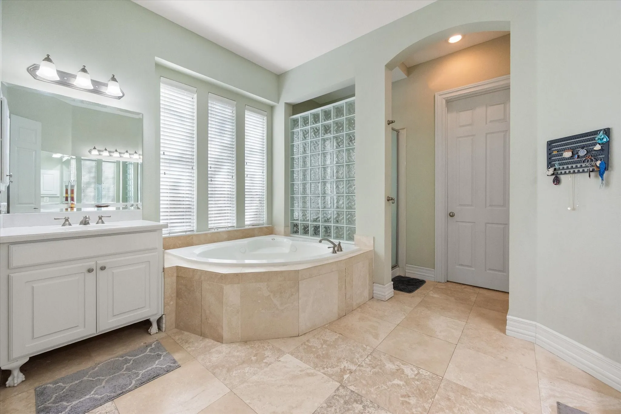  Luxurious primary bathroom retreat featuring a relaxing jetted soaking tub surrounded by elegant travertine tile, perfect for unwinding after a long day. Spacious layout includes a separate walk-in shower and a stylish vanity area with furniture-style cabinetry. Large windows and glass block accents provide abundant natural light while maintaining privacy. Beautiful travertine flooring throughout adds warmth and sophistication, creating a spa-like atmosphere designed for comfort and functionality.