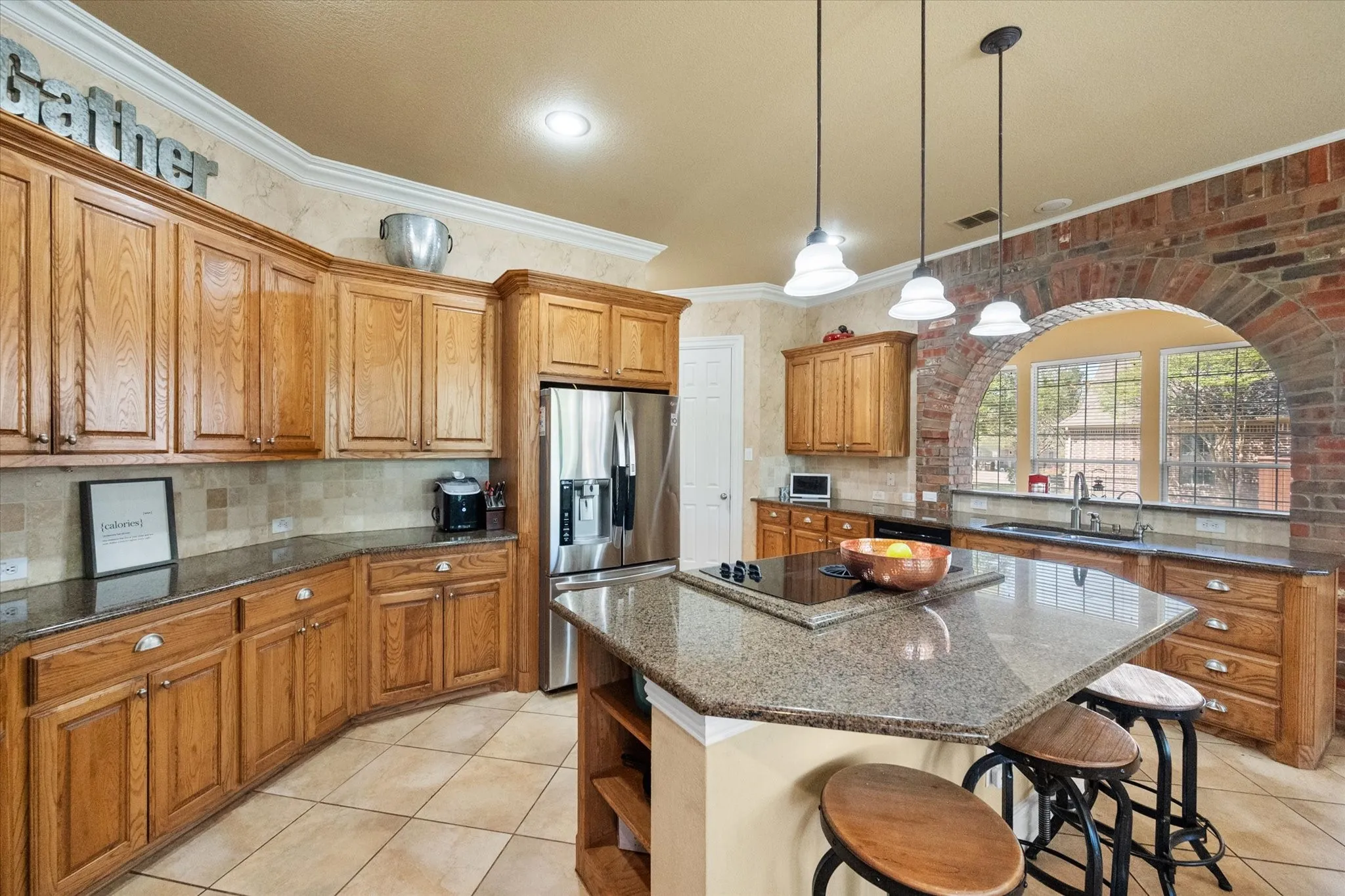 Gourmet kitchen featuring a wall of custom cabinetry offering abundant storage, perfect for organization and functionality. Expansive center island with built-in cooktop and oversized breakfast bar creates the ideal space for entertaining and everyday living. Granite countertops, stainless steel appliances, and warm neutral tones complement the space, while pendant lighting adds style and ambiance. Open layout with tile flooring, ample counter space, and natural light makes this kitchen both inviting and highly functional—perfect for hosting and family gatherings.