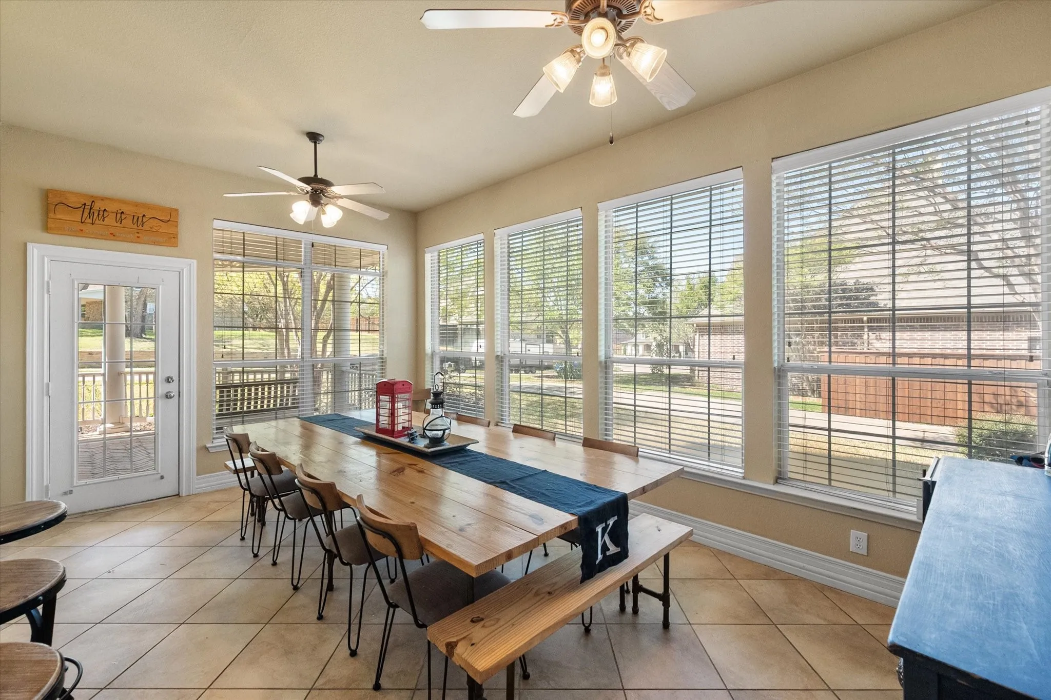Versatile sunroom dining space filled with natural light, featuring a wall of oversized windows and serene backyard views. Currently styled as a spacious dining area, this flexible room easily accommodates a large table seating 10—perfect for holiday dinners, entertaining, or large family gatherings. Neutral tones, tile flooring, and dual ceiling fans enhance comfort and functionality, while direct outdoor access adds convenience. Ideal as a sunroom, second dining area, or multi-purpose living space designed for both everyday living and special occasions.