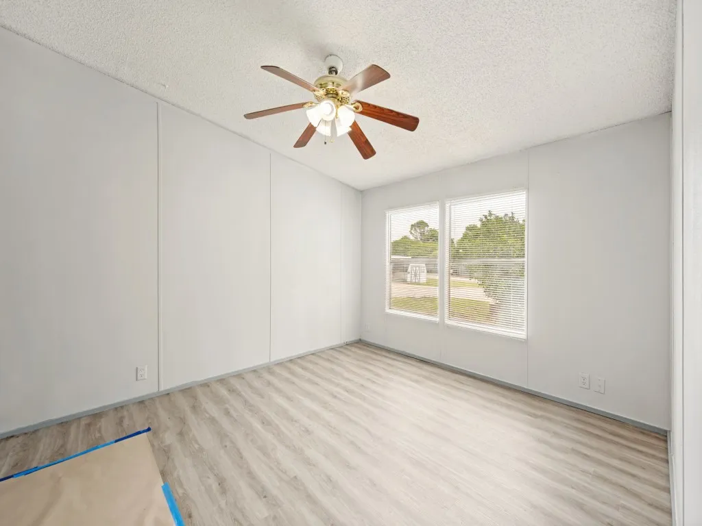 Spacious room featuring wood-finish flooring, two large windows, a ceiling fan with integrated lighting, and neutral wall paneling
