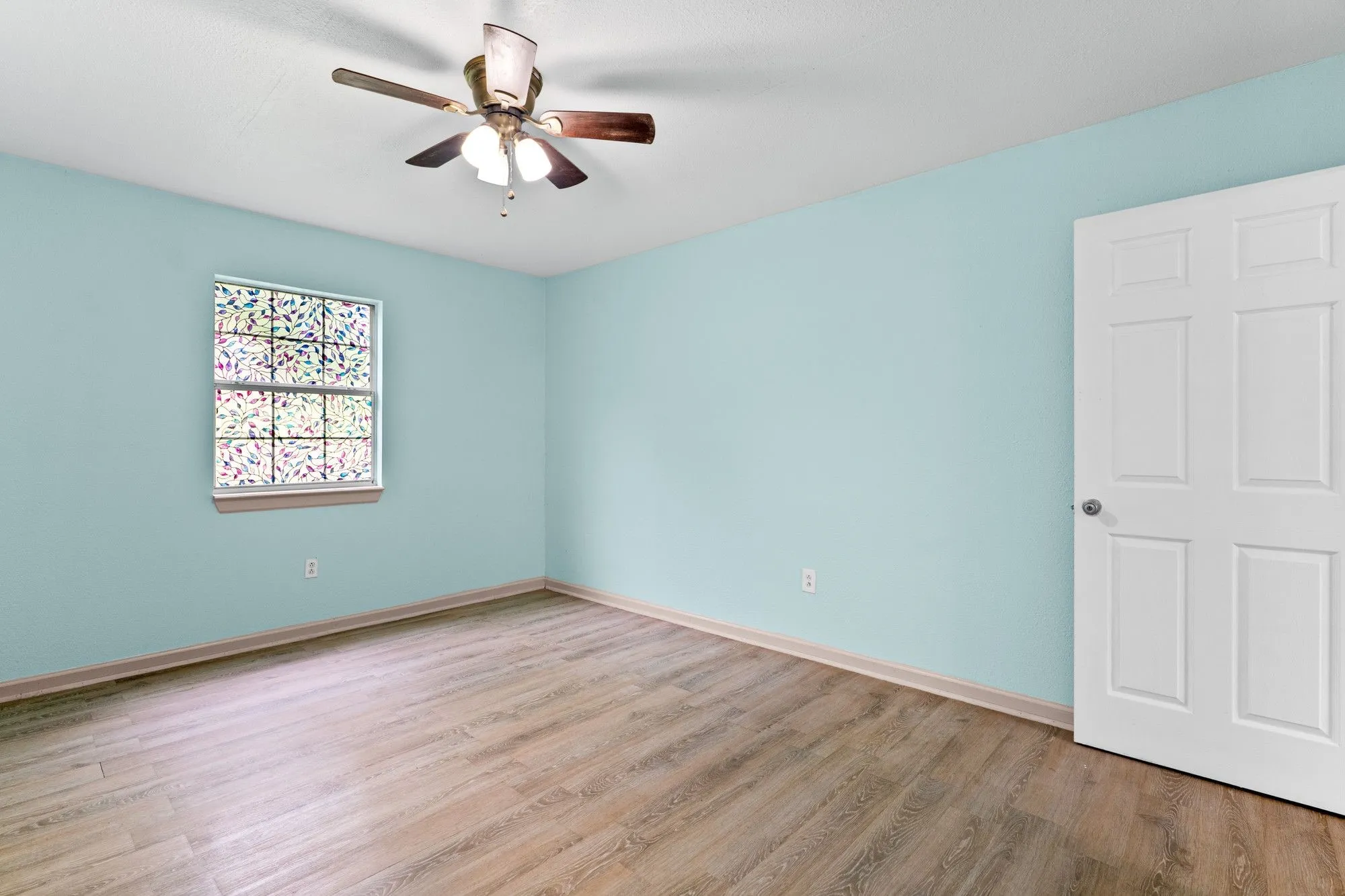 Room featuring wood-finish flooring, light blue wall paint, a multi-pane window with privacy film, a white paneled door, and a ceiling fan with light fixture